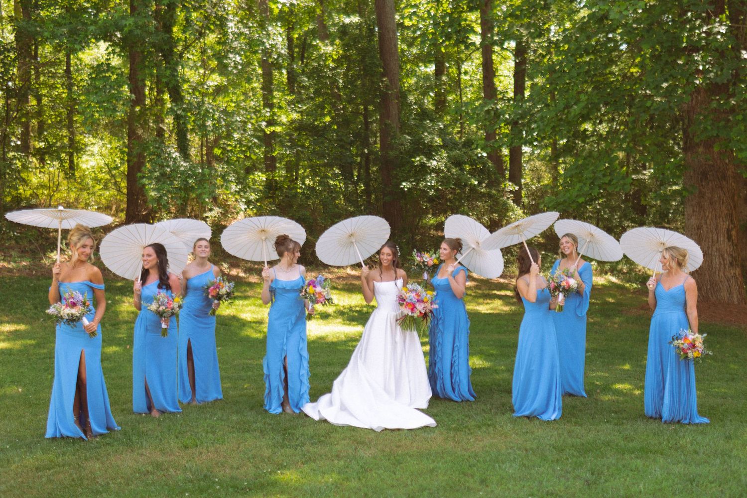 Bridesmaids in blue gowns holding white parasols pose with bride in lush green outdoor setting.