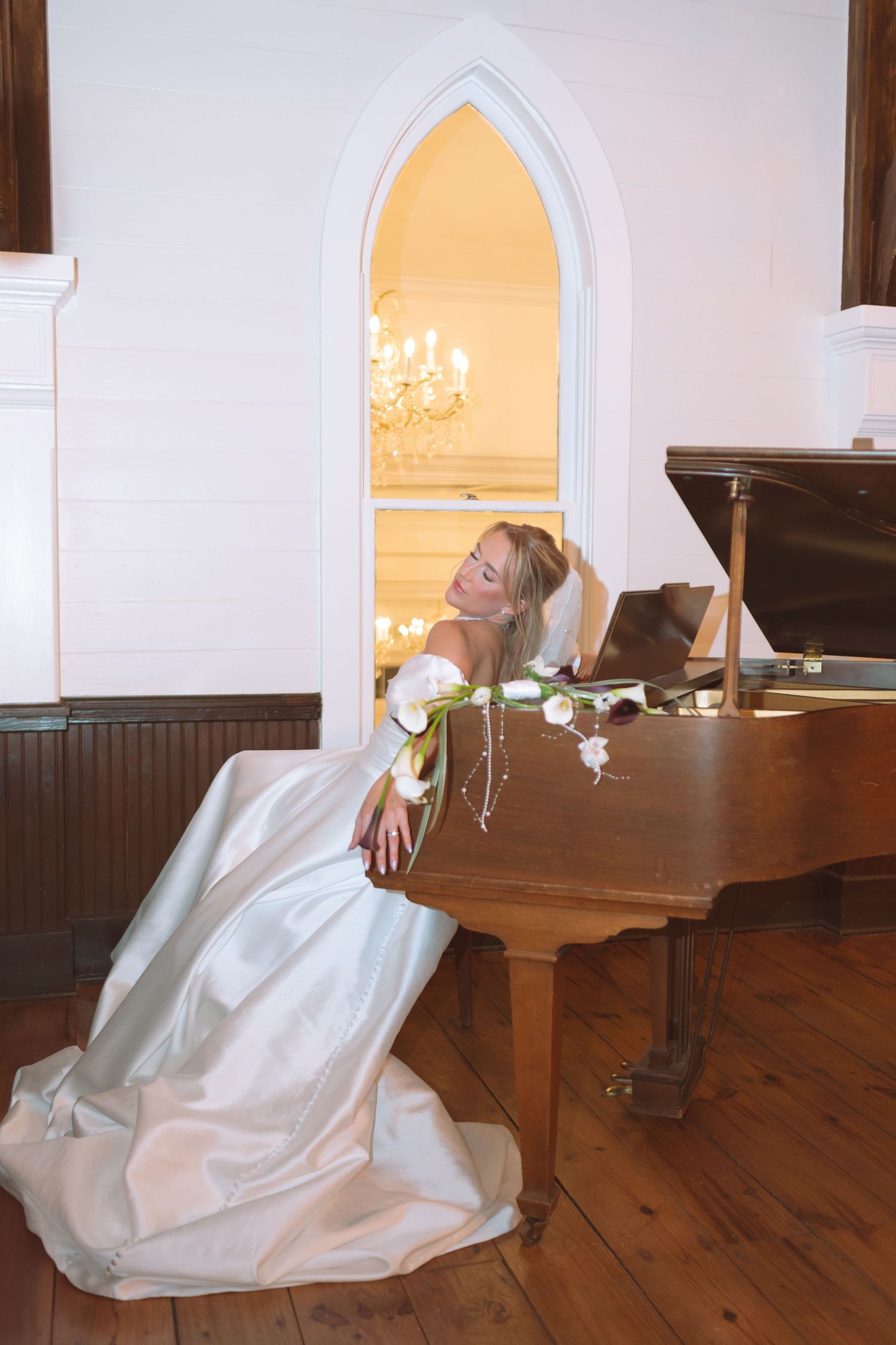 Bride in white gown posing gracefully beside a grand piano in a chapel.