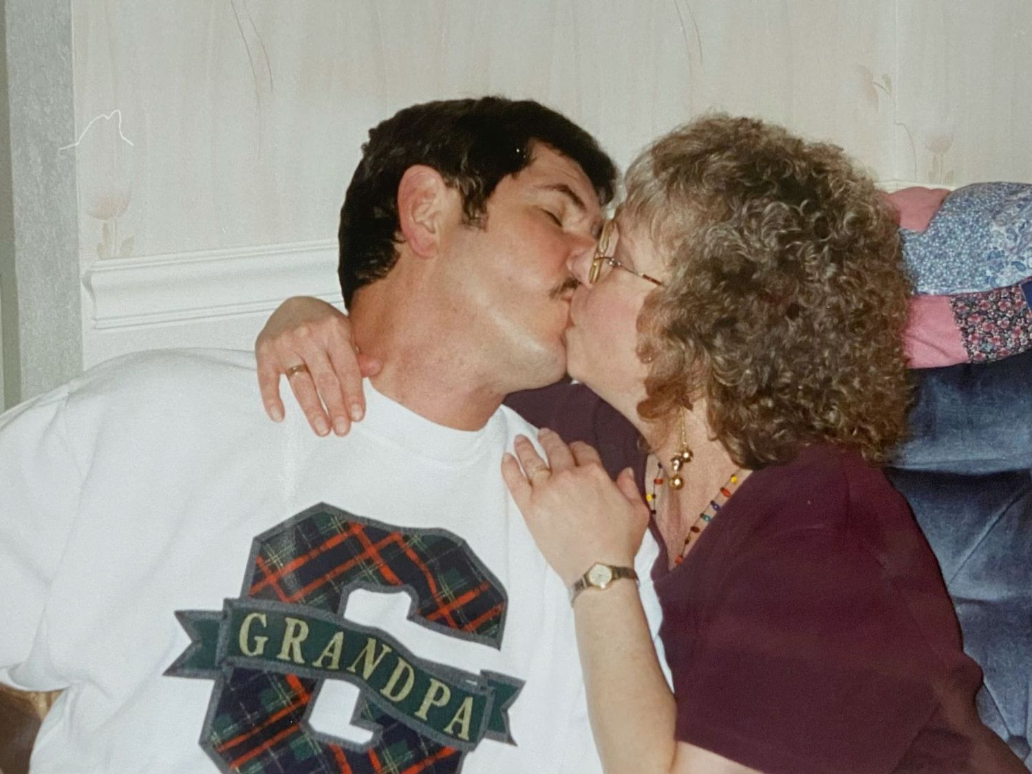 Man wearing Grandpa shirt kissing a woman with curly hair indoors.