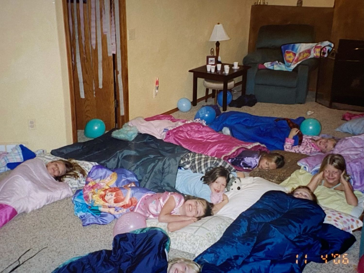 Girls sleeping in sleeping bags on the floor at a slumber party with teal balloons decorating the room.