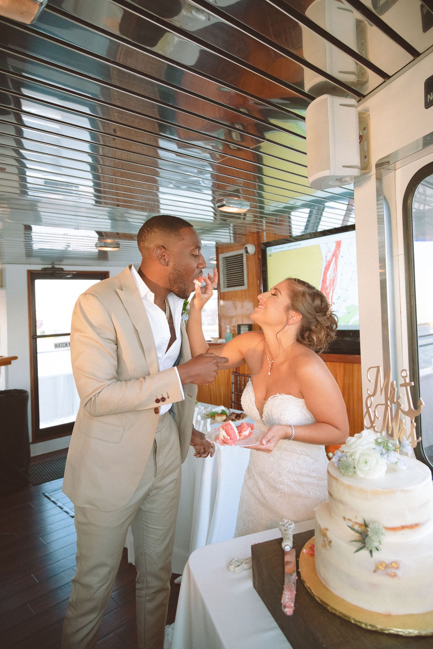 Interracial couple exchanges cake during wedding reception aboard a yacht with wooden interior decor.