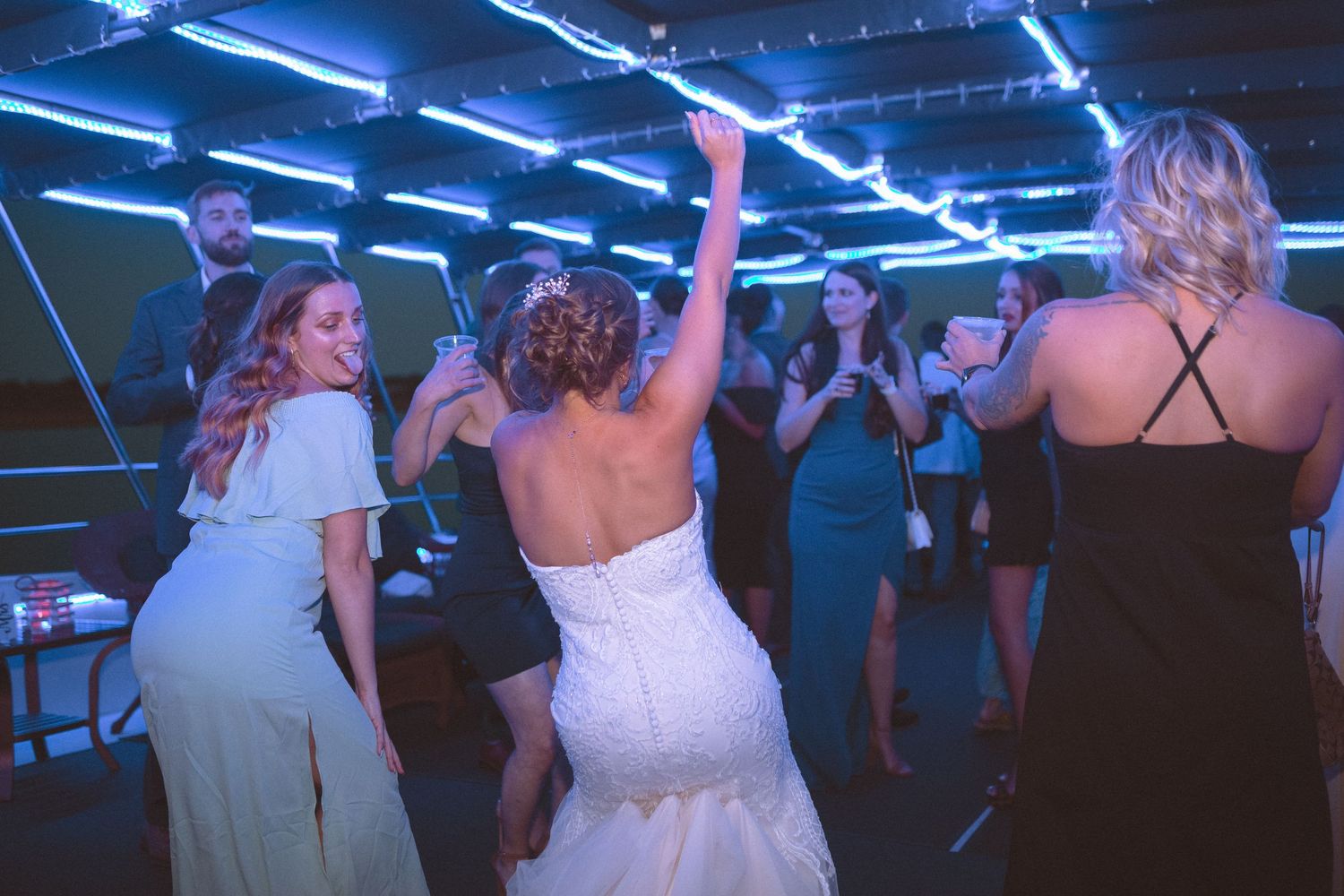 Bride in white wedding gown dances with guests under blue neon lights at wedding reception.