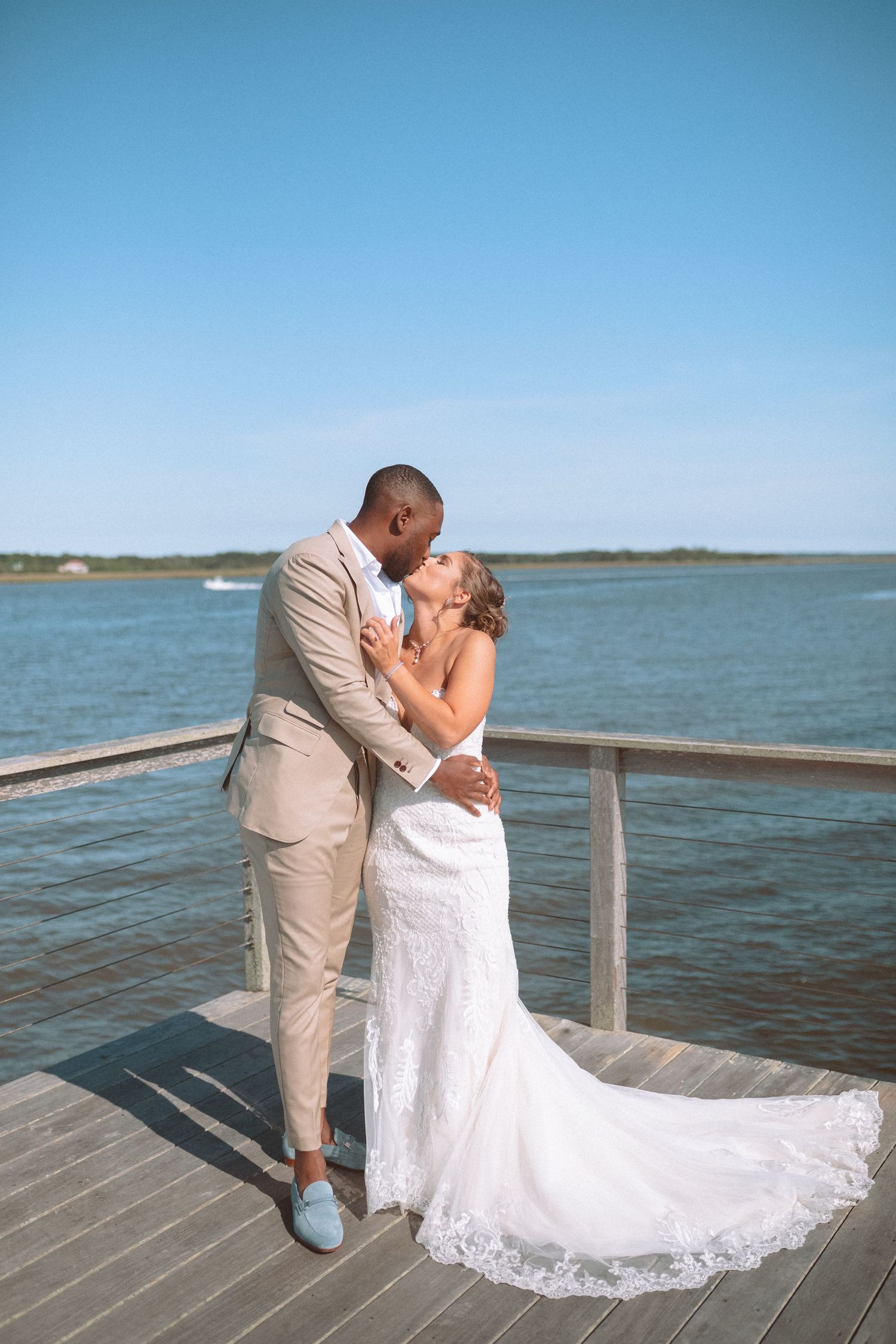 Interracial couple sharing a kiss on a waterfront dock in wedding attire, bride in lace gown with train.