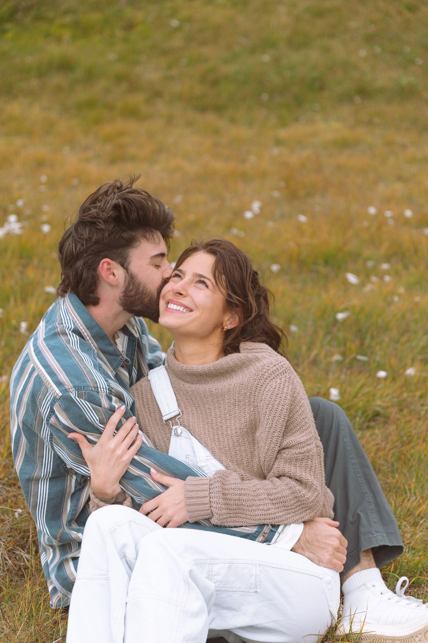 Happy couple sitting on grass, man kissing smiling woman on cheek in meadow.