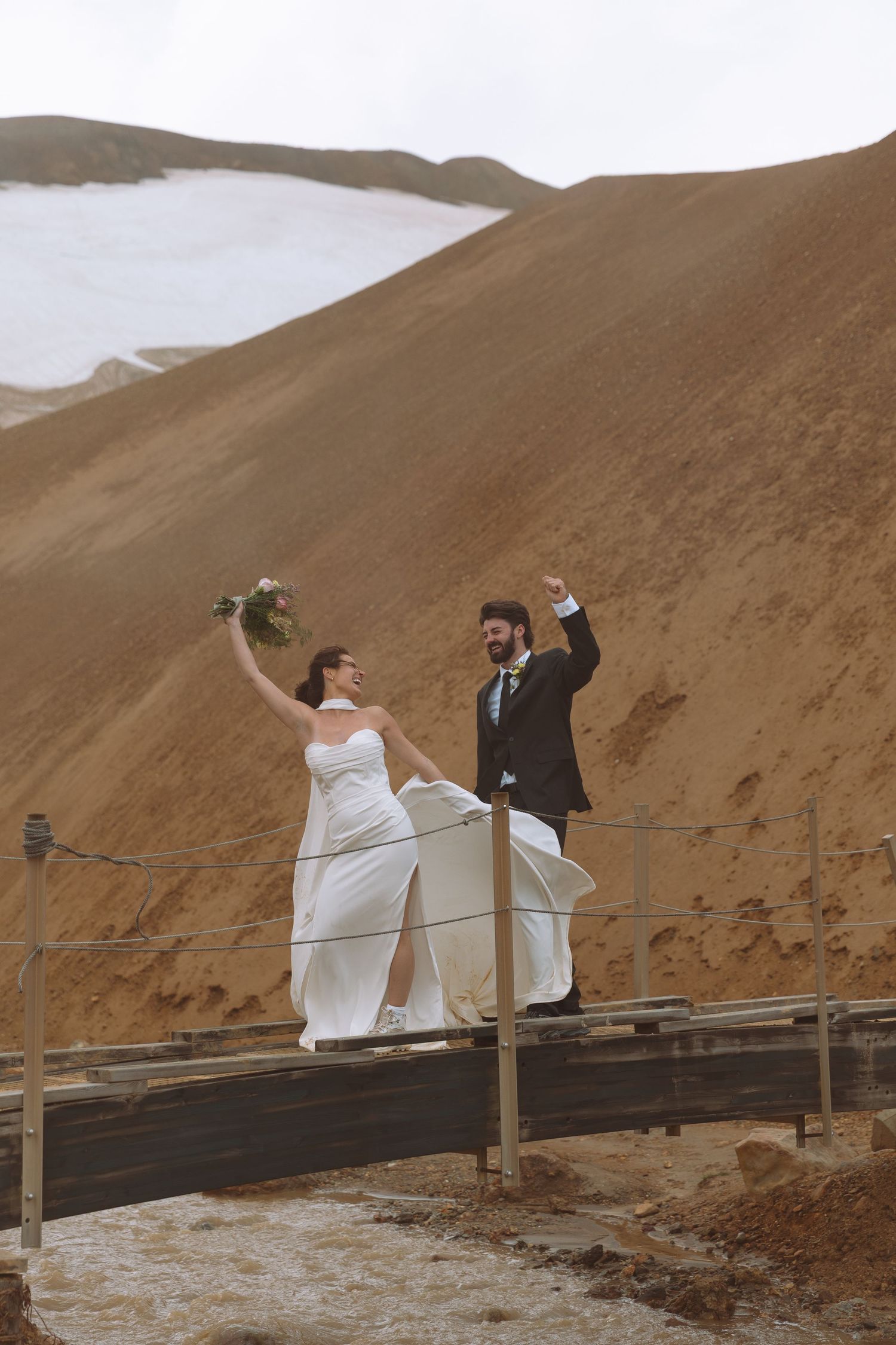 Joyful bride and groom celebrate with raised arms on a wooden bridge in Iceland highland landscape.