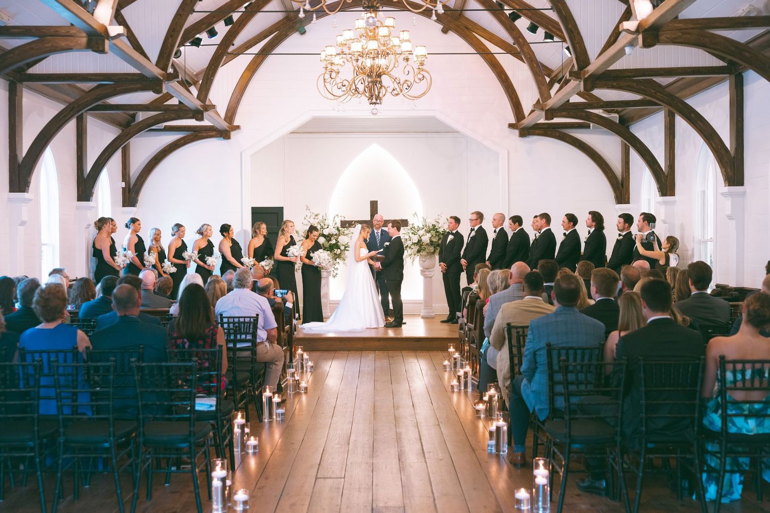 Romantic wedding ceremony in a white chapel with wooden beams, candlelit aisle, and guests seated in dark chiavari chairs.