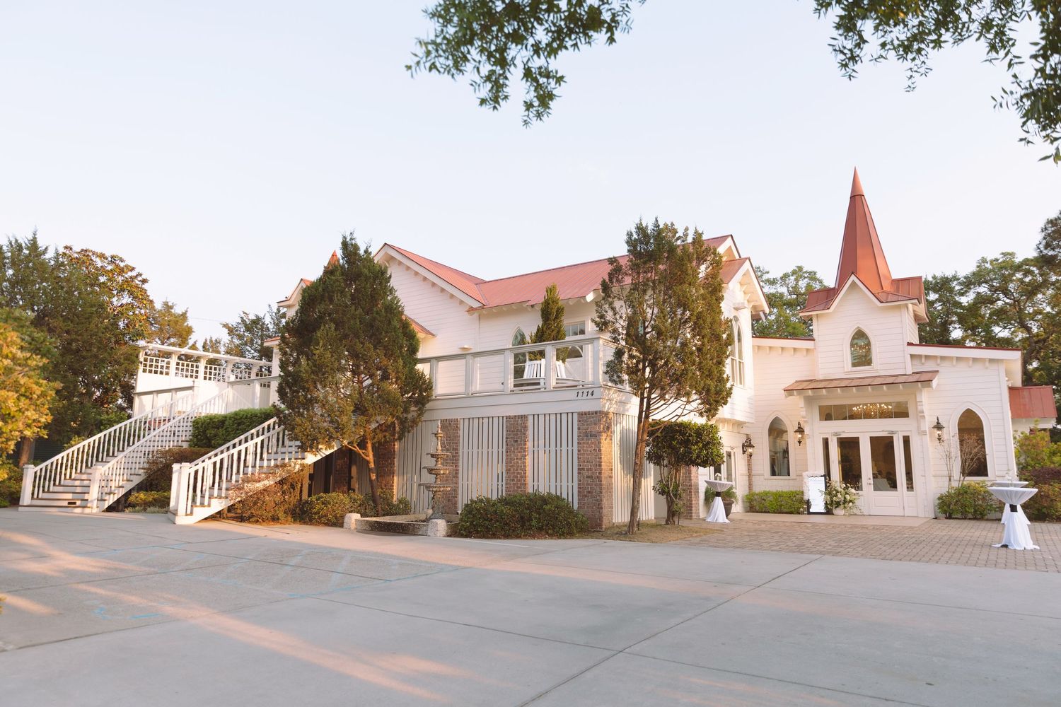Elegant white wedding venue with red roof chapel, grand staircase, and lush trees in warm evening light.