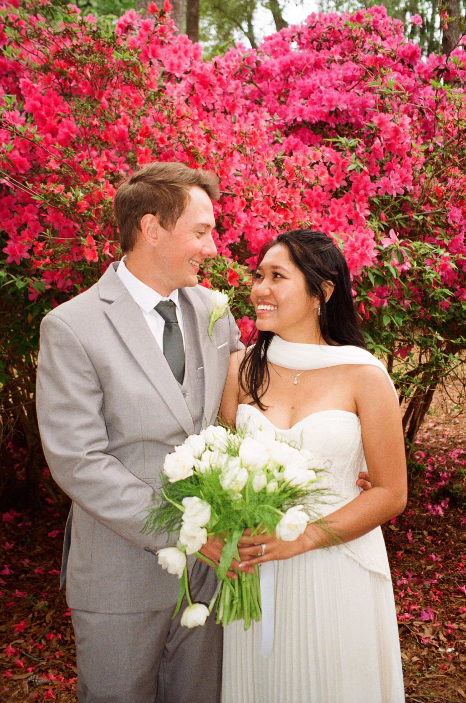 Bride and groom smiling together in front of vibrant pink azalea bushes, bride holding white tulip bouquet.