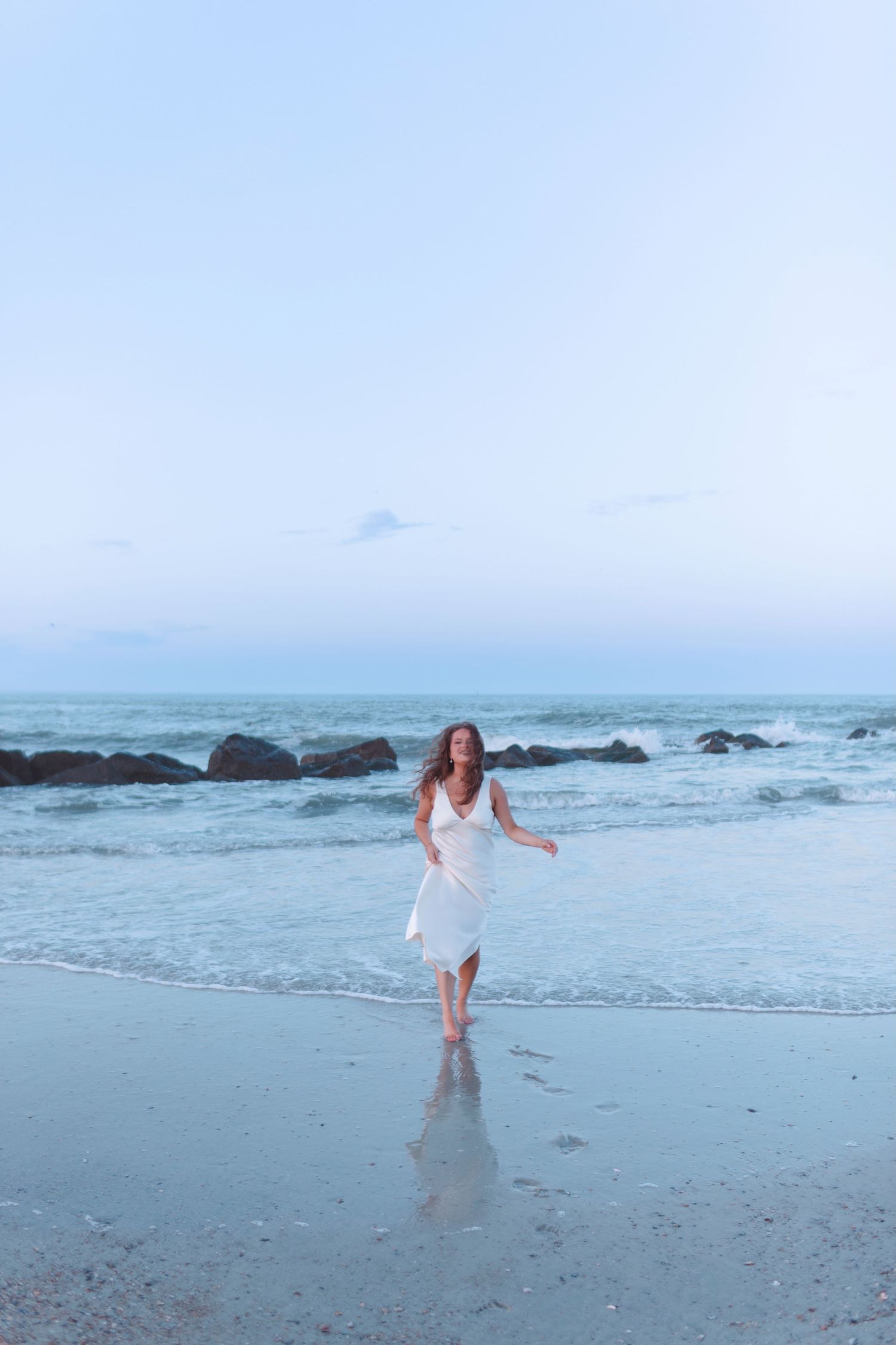 Woman in white sundress walking along shoreline at twilight with rocky ocean backdrop.