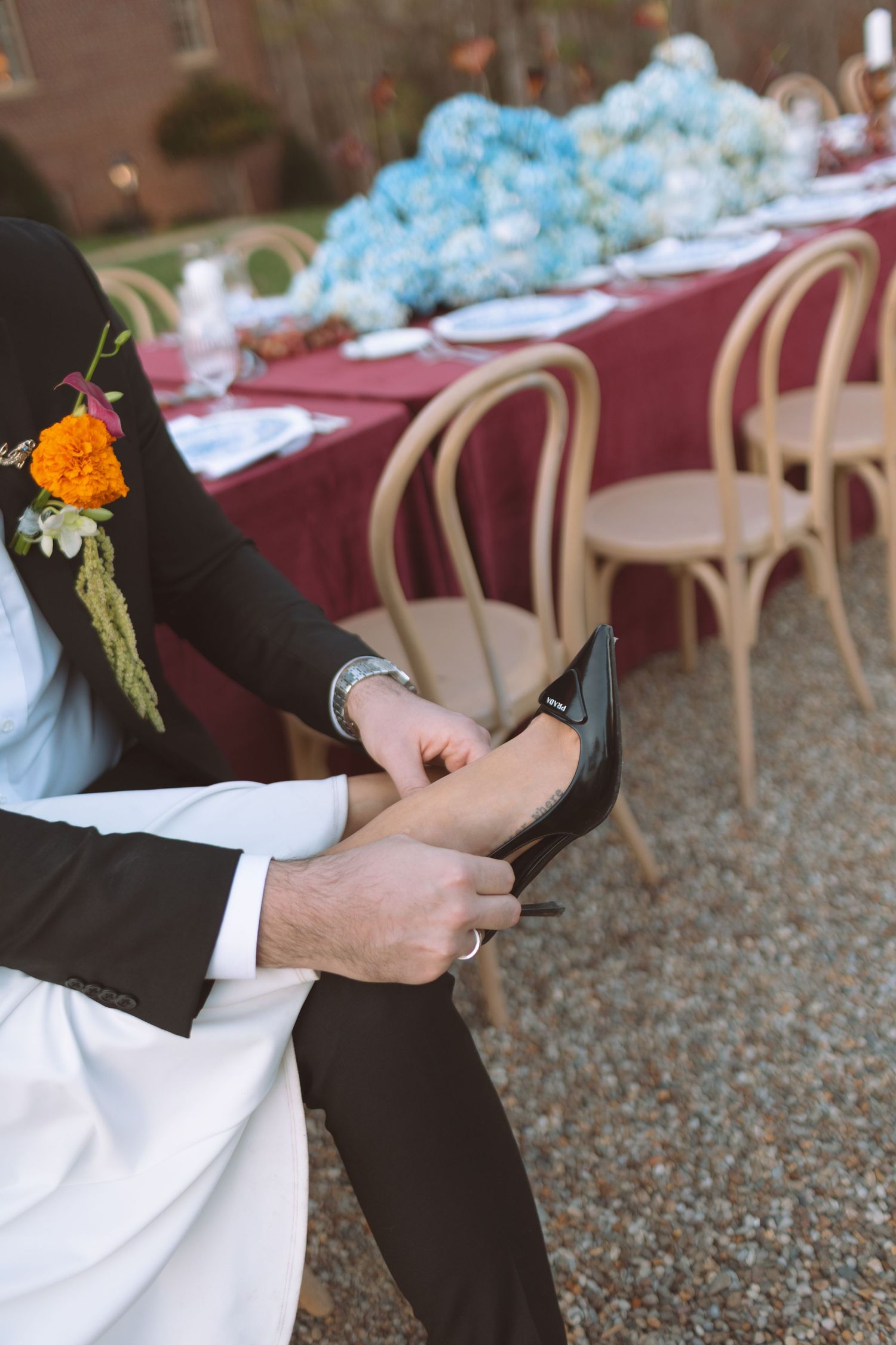 Groom helping bride put on a black high heel shoe beside an elegantly decorated outdoor reception table with blue florals.