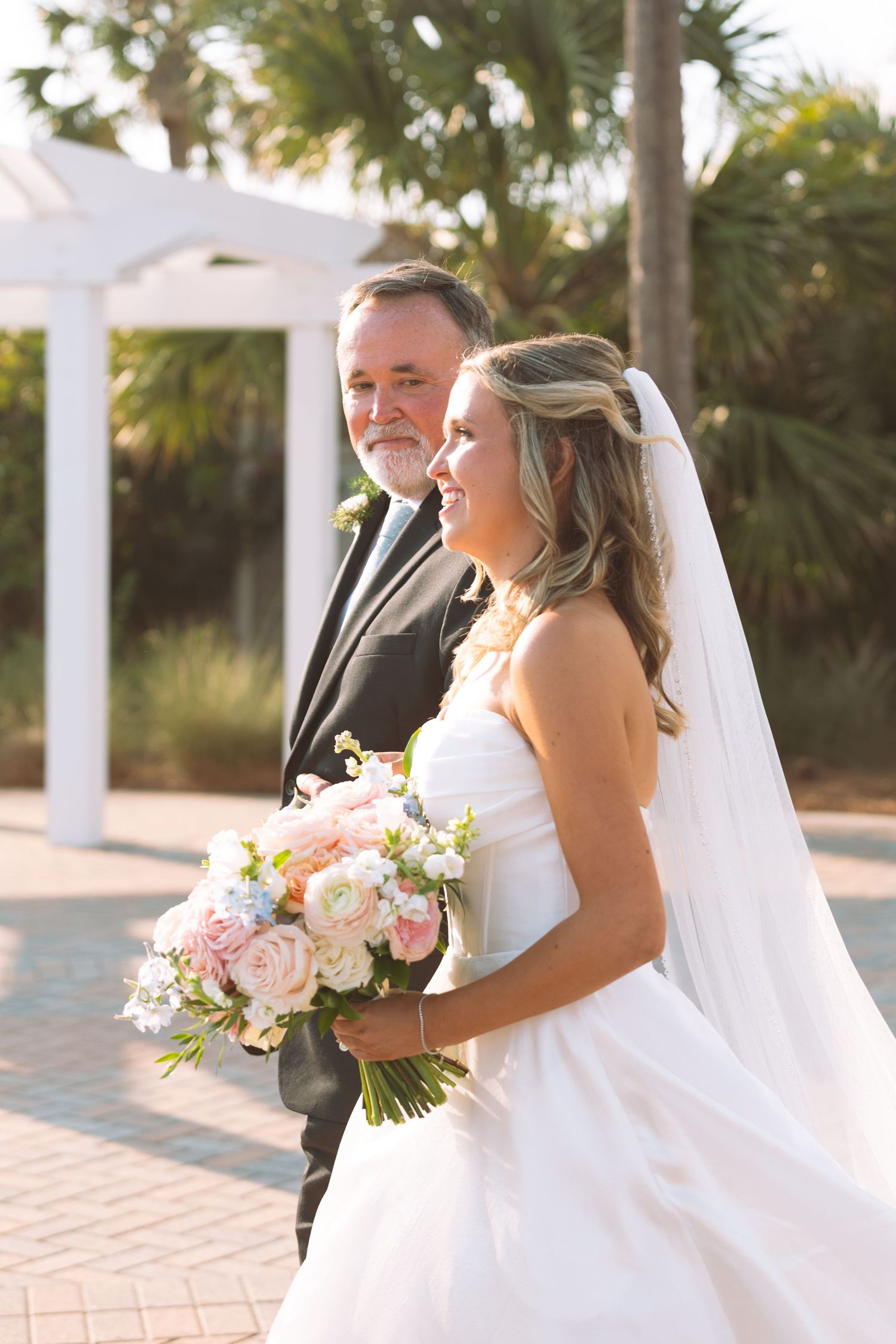 Smiling bride in white strapless gown holding pink bouquet walking with older gentleman near palm trees.