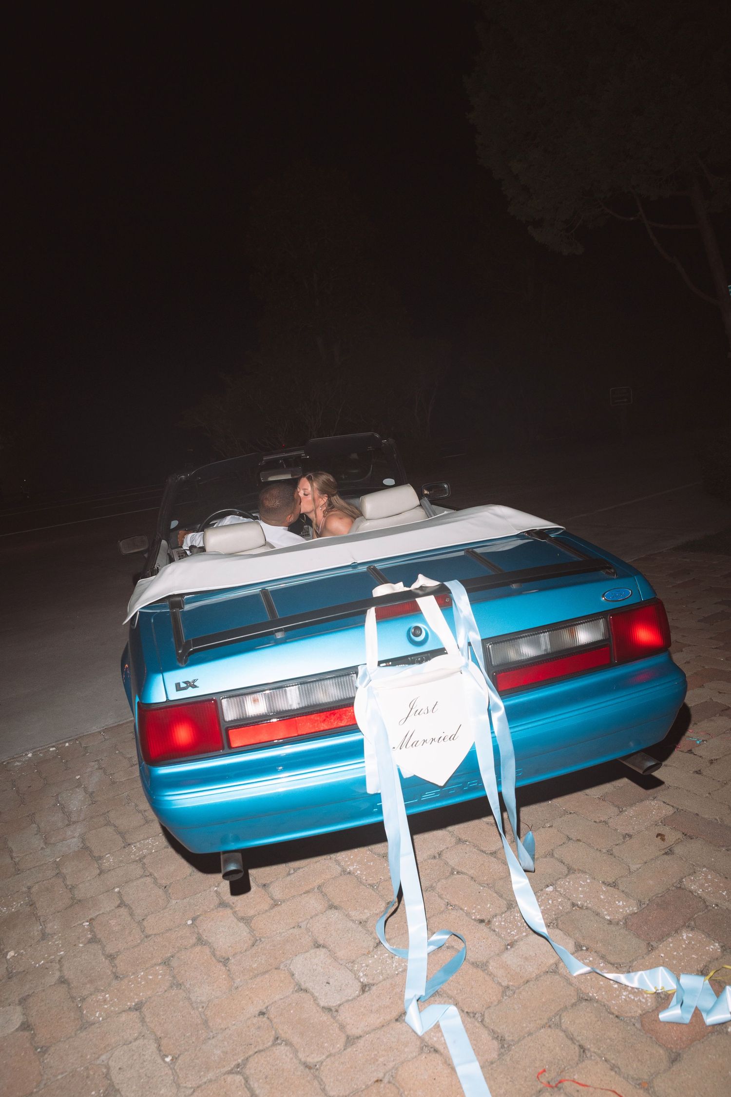 Newlyweds driving away in a blue convertible decorated with a Just Married sign at night.