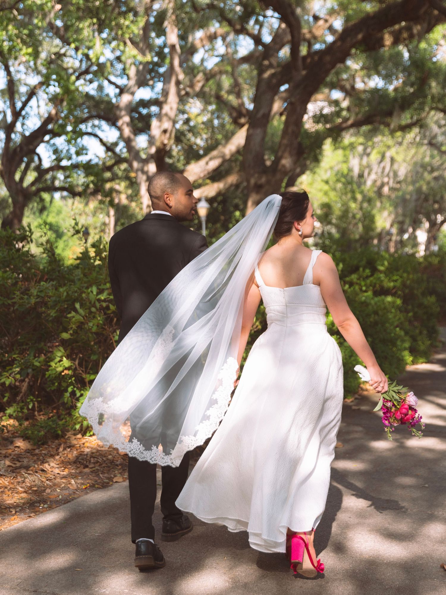 Bride and groom walking hand in hand under oak trees, bride wearing pink heels and flowing veil.
