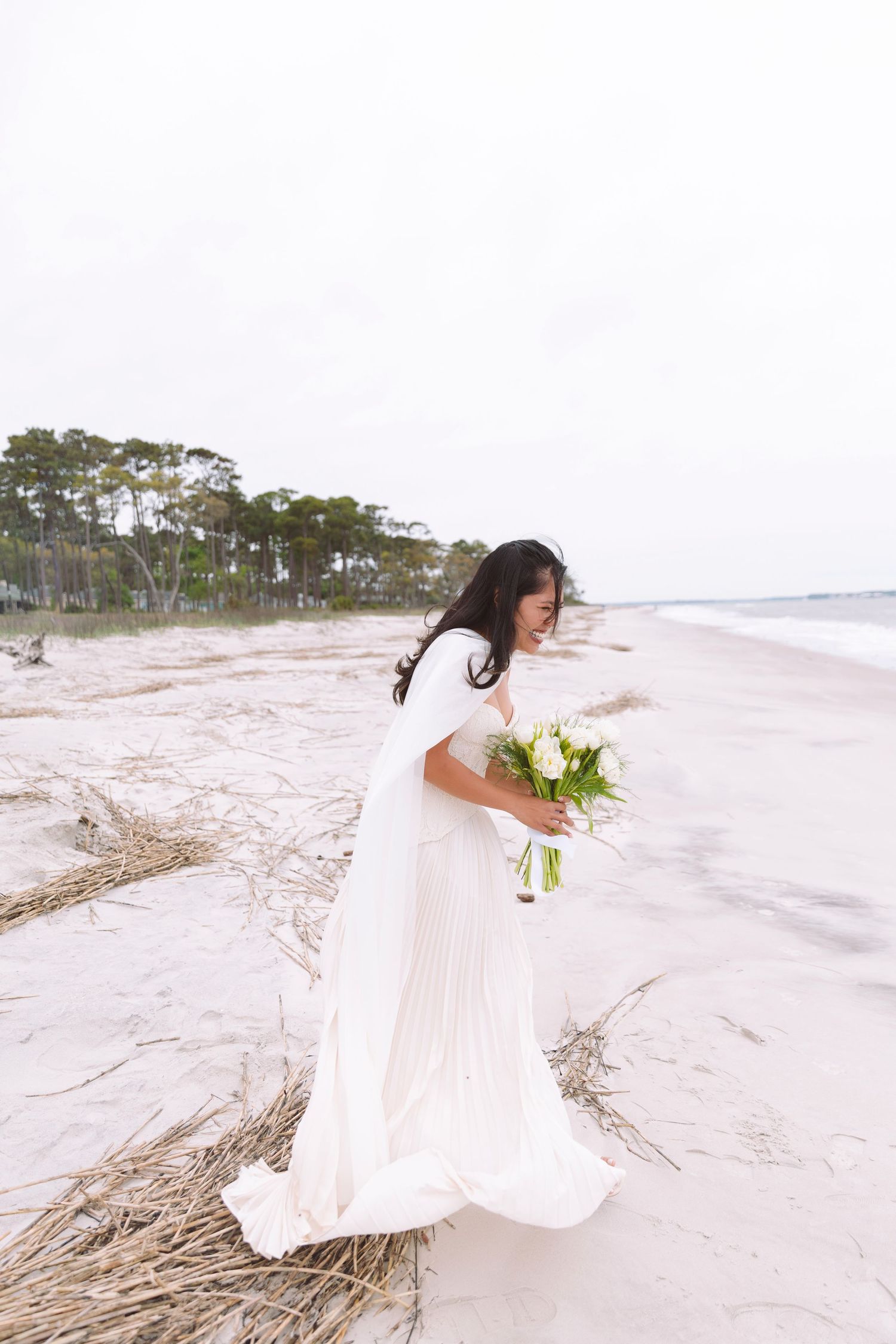 Bride in a flowing white gown holding a white bouquet, standing on a windy beach with pine trees behind.
