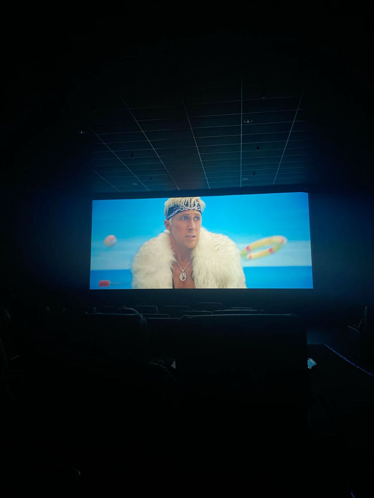 Movie theater screen showing a man in a white fur coat and blue headband in a bright scene.