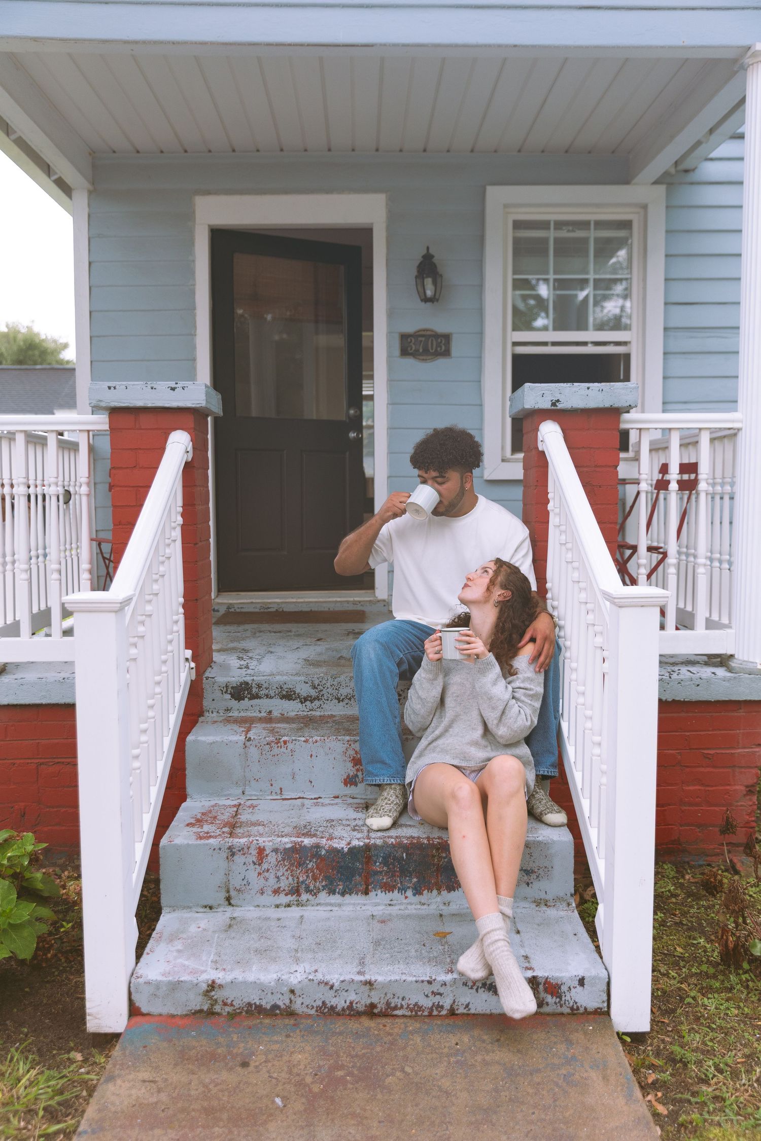 Couple sitting on front porch steps of a blue house drinking coffee together.