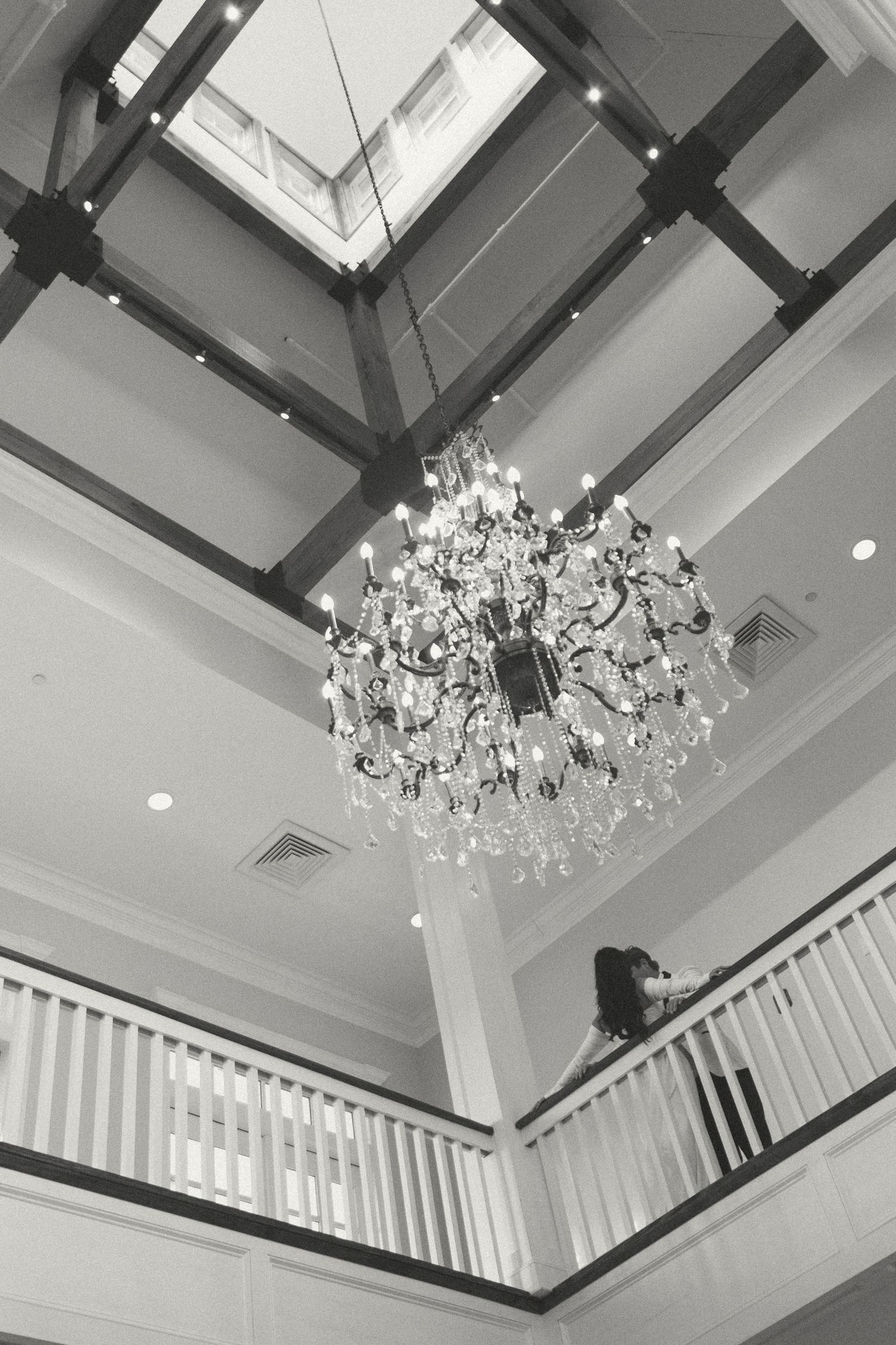 Black and white low-angle shot of a grand crystal chandelier with a bride visible on the balcony below a skylight.
