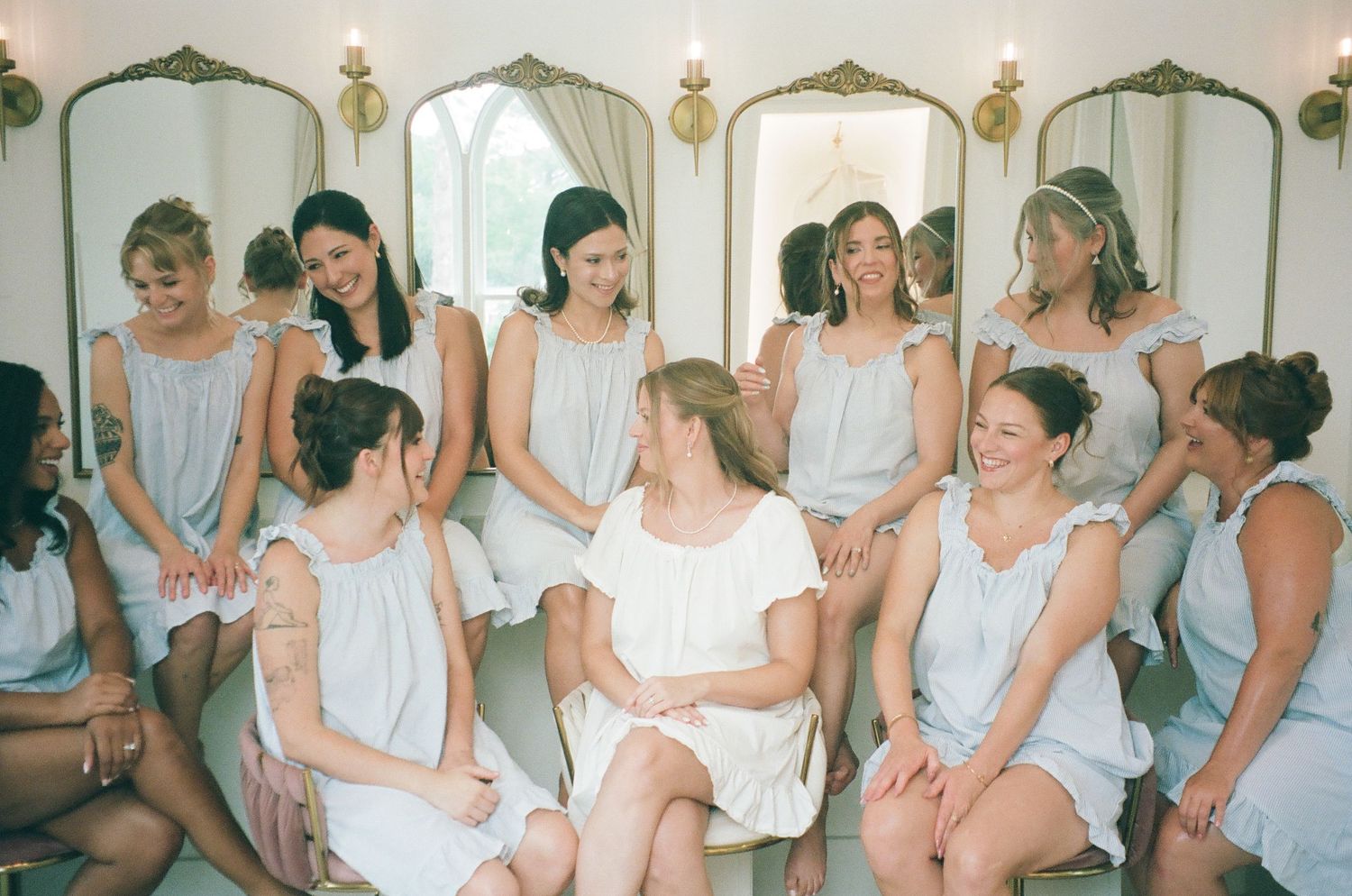 Bridesmaids in white dresses sit together laughing in an elegant room with gold mirrors during wedding preparations.
