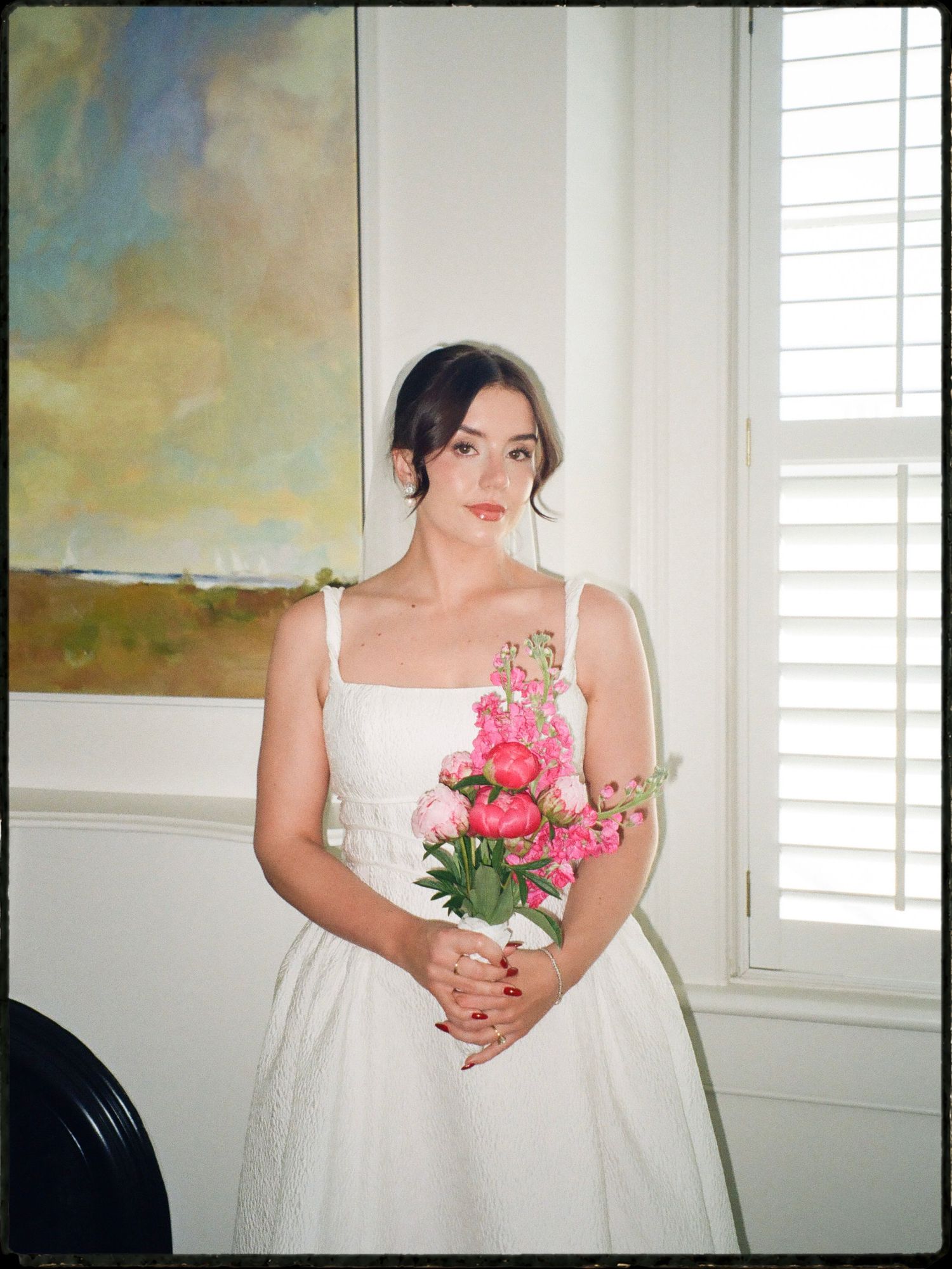Bride in white square-neck dress holding pink peony bouquet beside window indoors.