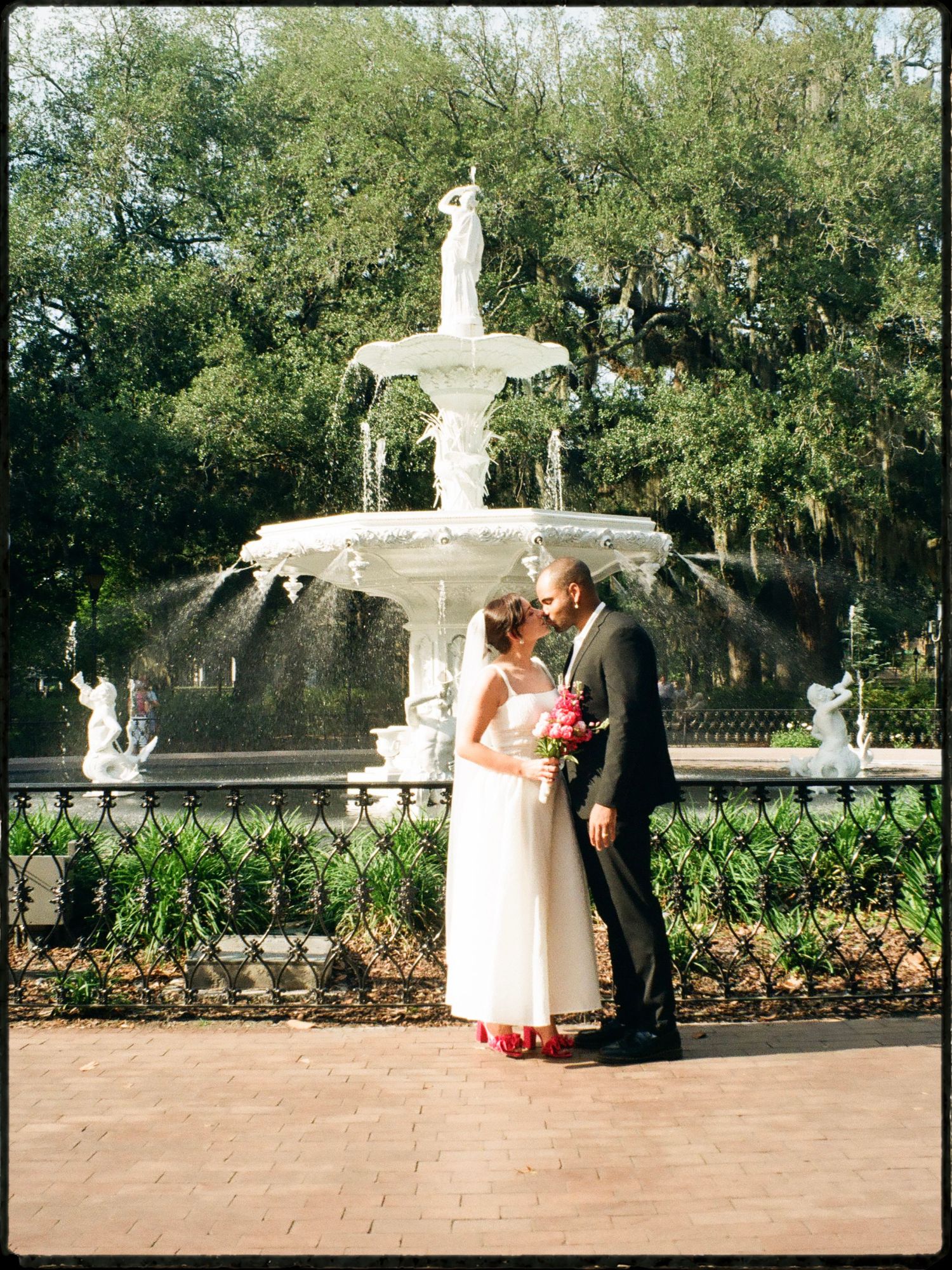 Bride and groom share romantic moment before elegant white tiered fountain in lush park setting.
