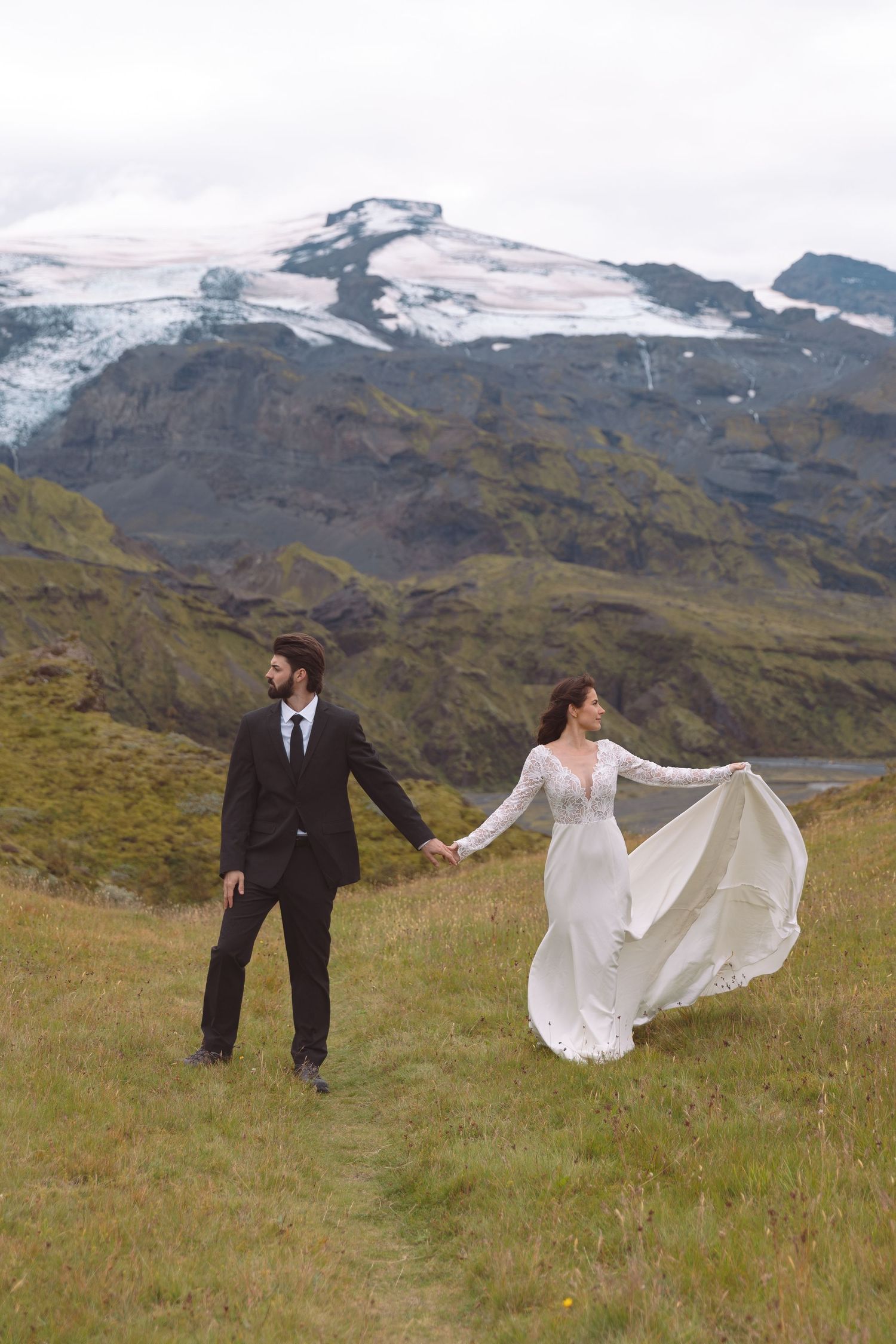 Bride and groom walk hand in hand through a mountain meadow, her white gown flowing in the wind.