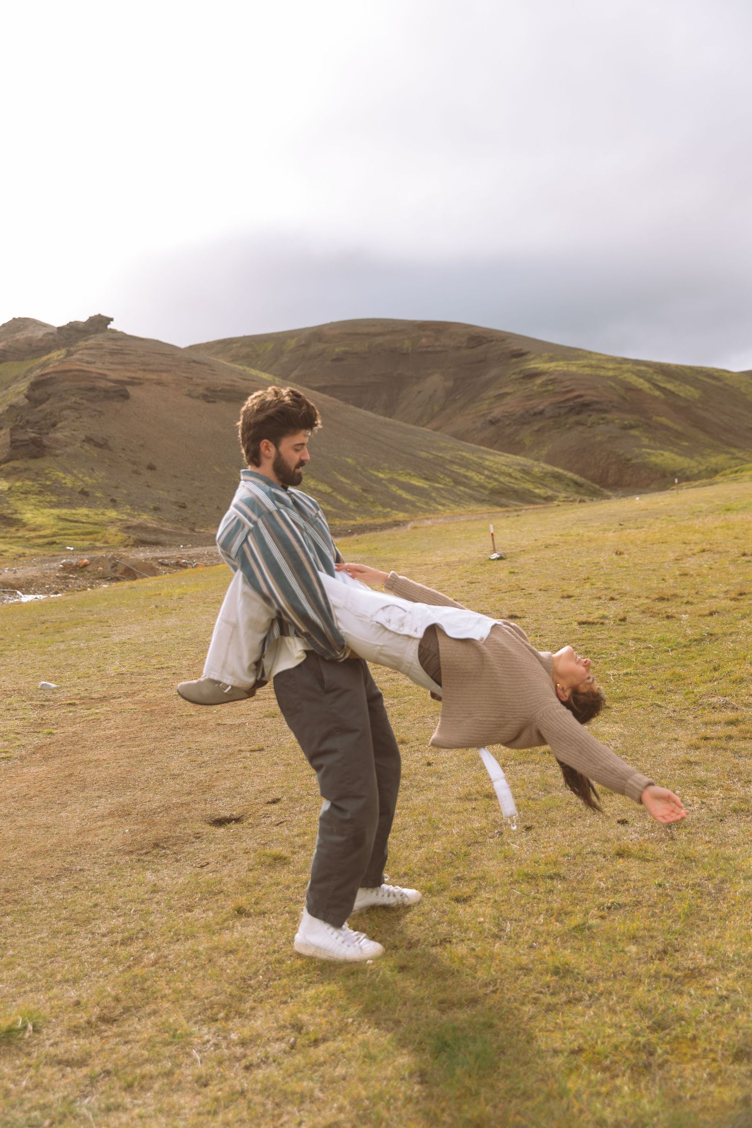 Man carrying laughing woman horizontally in a dramatic green mountain landscape under cloudy skies.