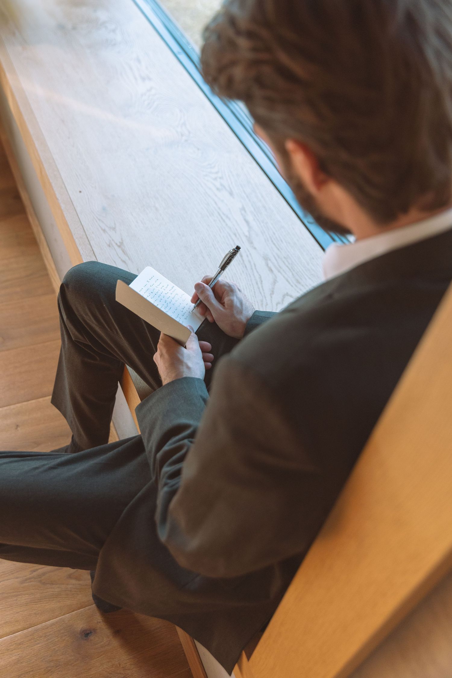 Aerial view of groom in dark suit writing personal wedding vows in notebook by window.