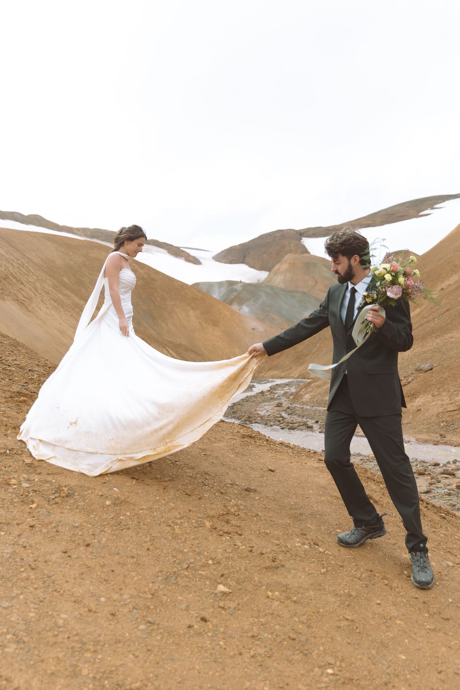 Groom holds bride's flowing white train while carrying a bouquet on a dramatic desert mountain landscape.