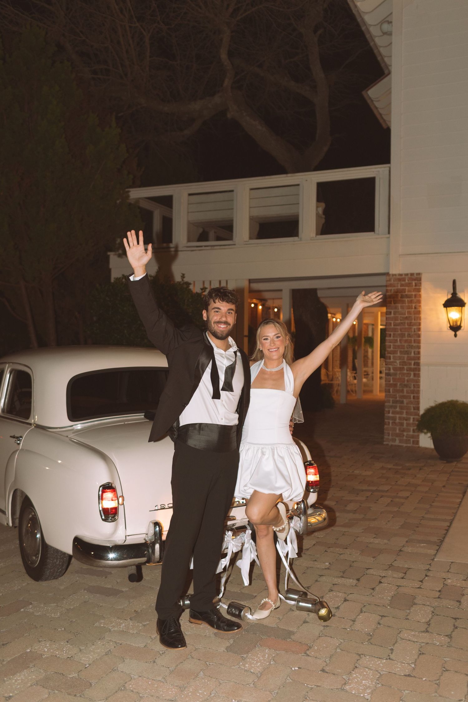 Joyful newlyweds waving beside vintage white classic car outside elegant wedding venue at night.