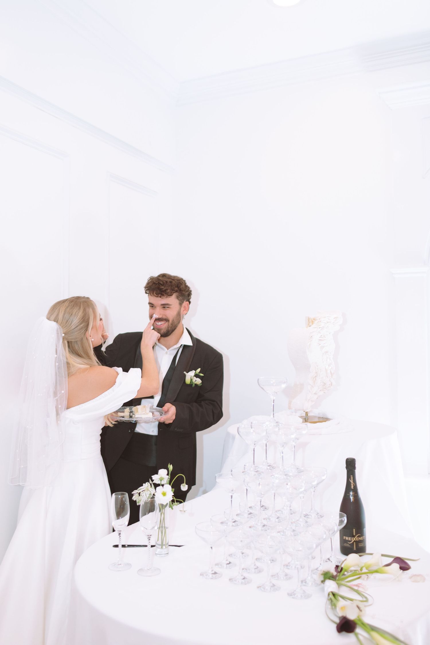 Bride feeding groom wedding cake beside a champagne coupe tower at their bright white reception venue.