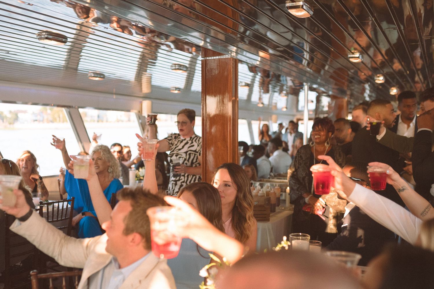 Lively wedding reception crowd raising glasses and dancing aboard a yacht with bright windows.