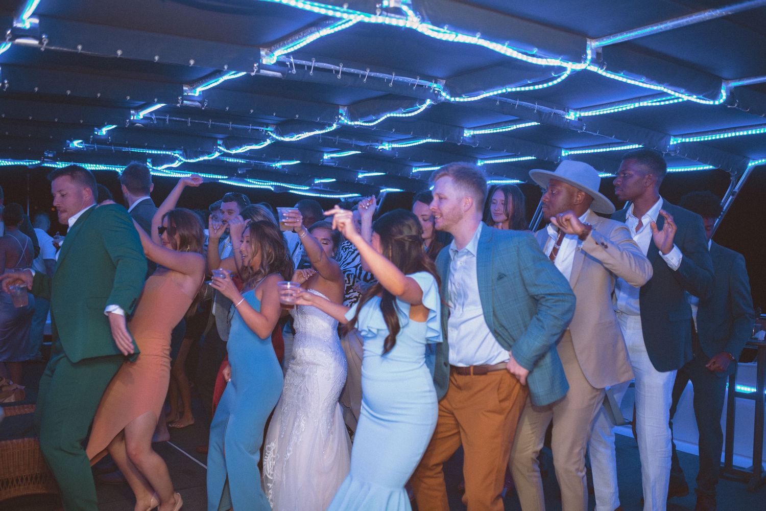 Wedding guests dancing joyfully at a lively reception party with blue neon lights overhead.