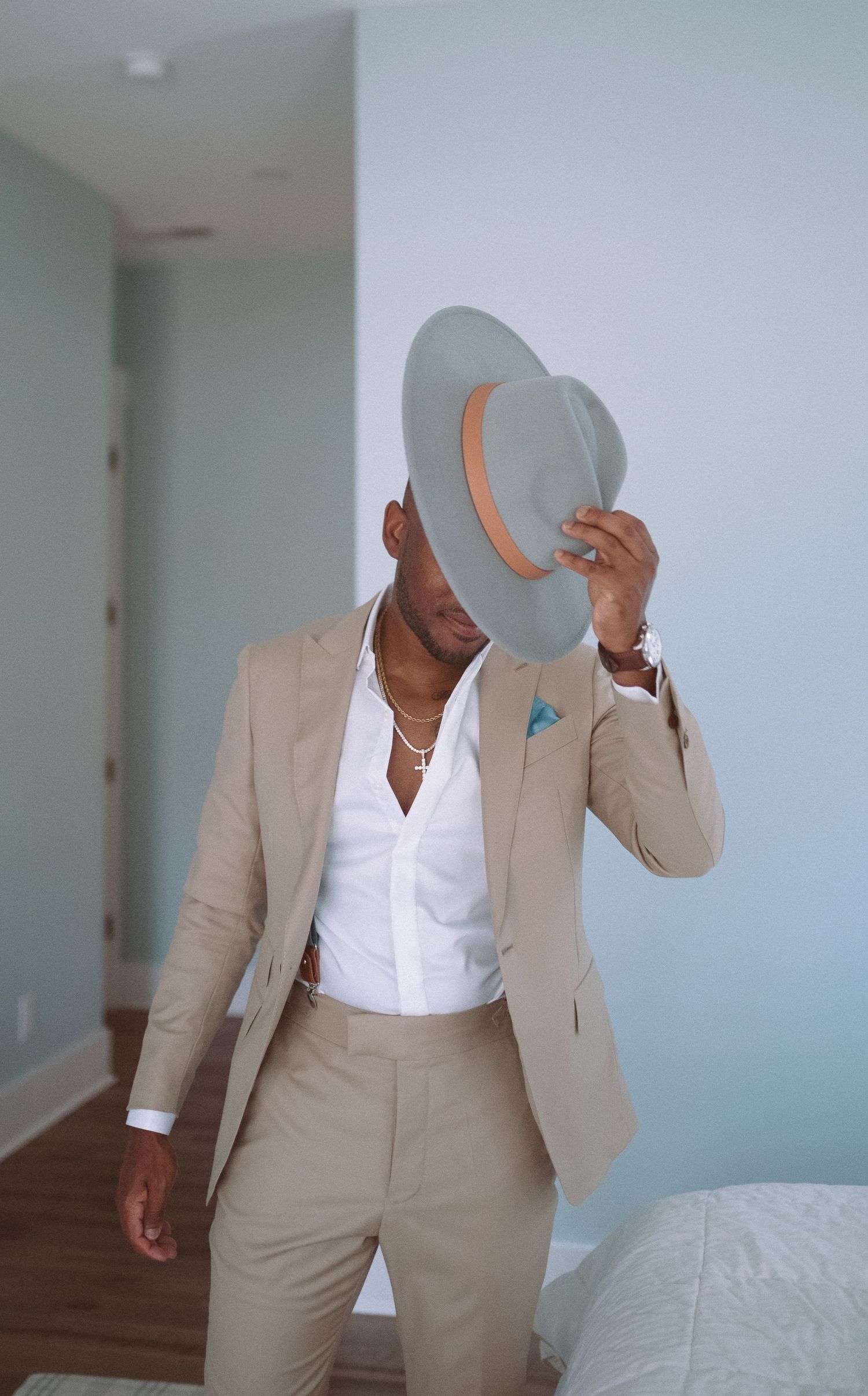 Stylish man in beige suit holding gray wide-brim hat over his face, wearing gold chain necklace and blue pocket square.