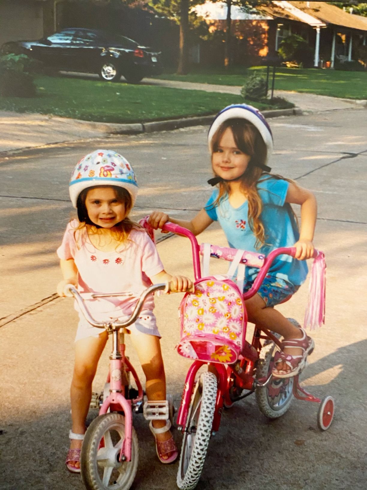 Two young girls smile while riding pink bikes with training wheels on a suburban street.