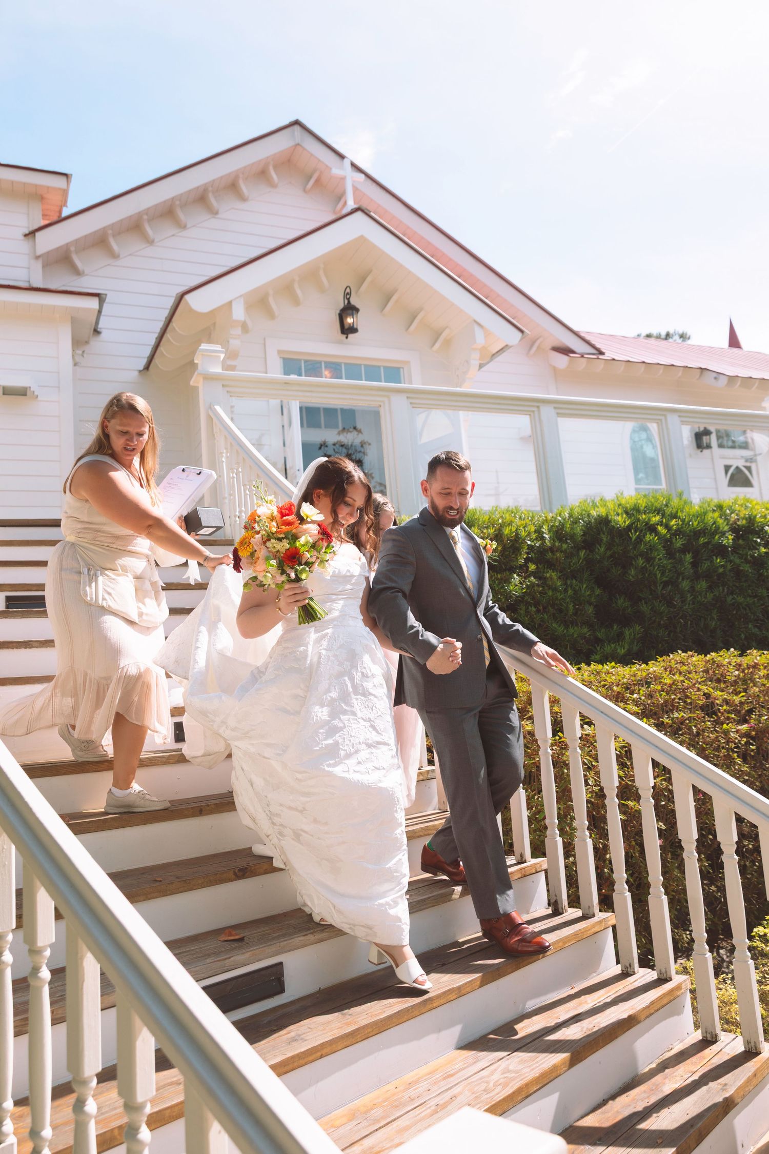Bride and groom descend white venue steps with bridesmaid, bride holding colorful bouquet on sunny wedding day.