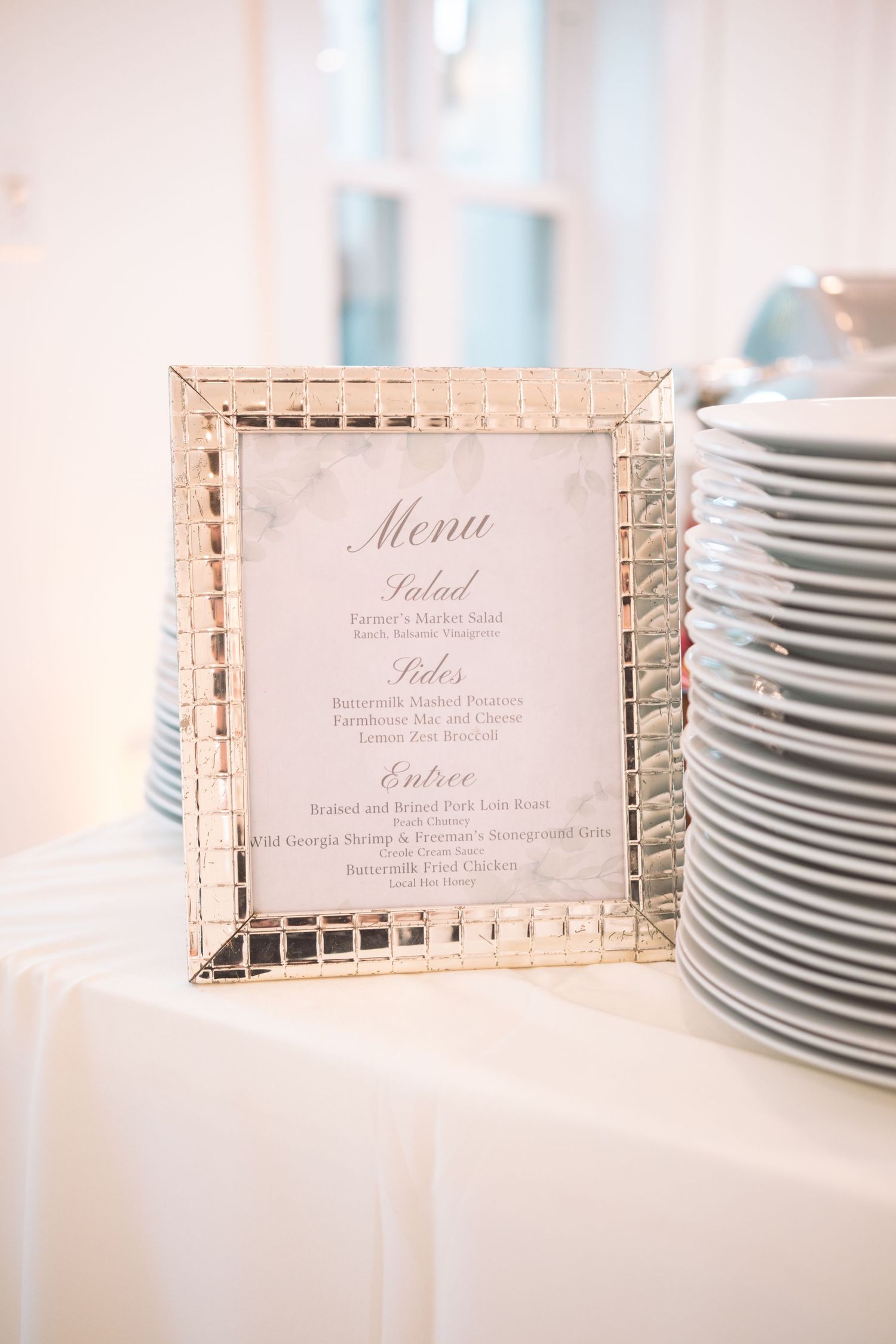 Elegant framed wedding menu card displayed beside stacked white plates at a reception dinner table.