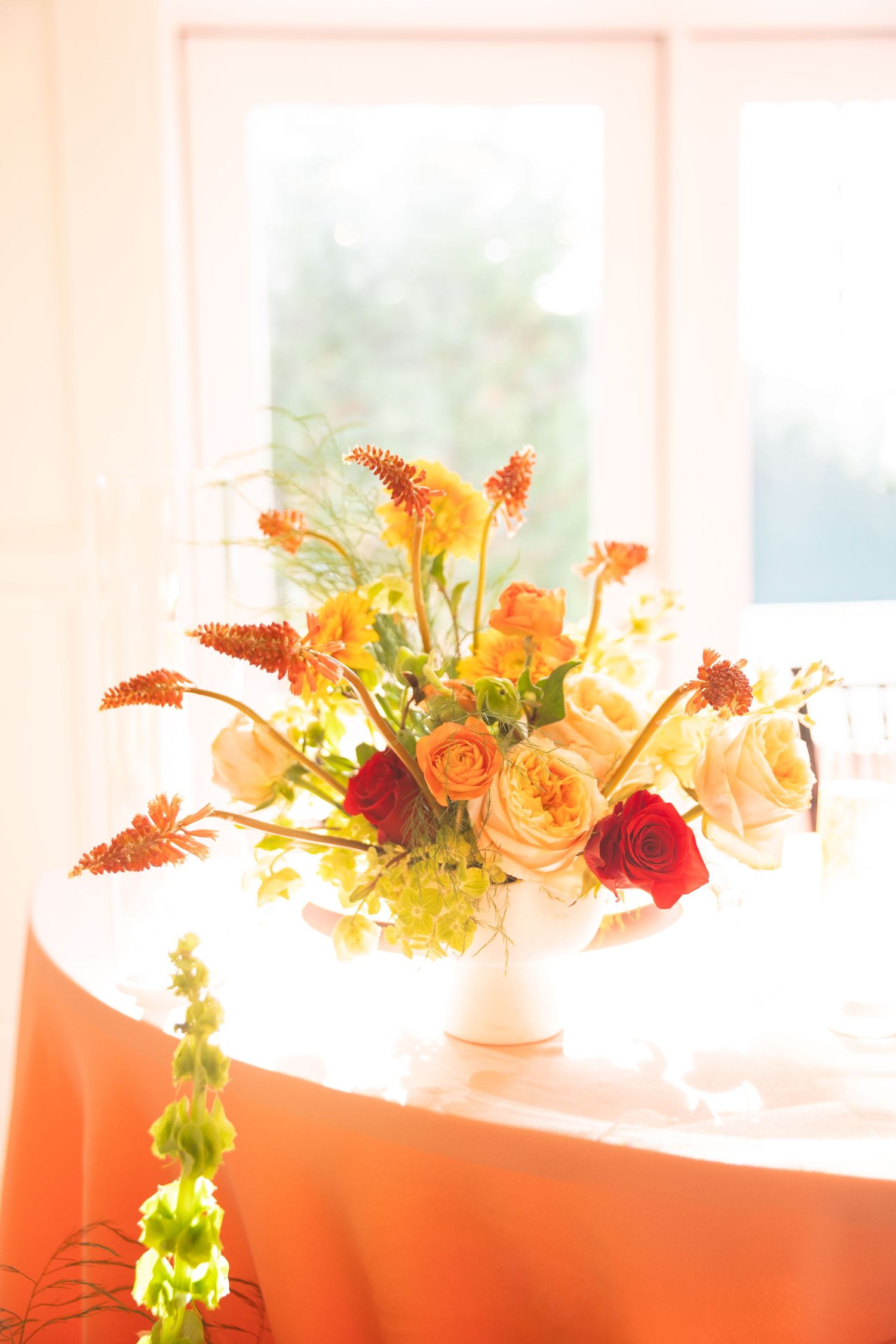 Elegant white vase with orange roses, red blooms, and wildflowers on an orange-draped table by a bright window.