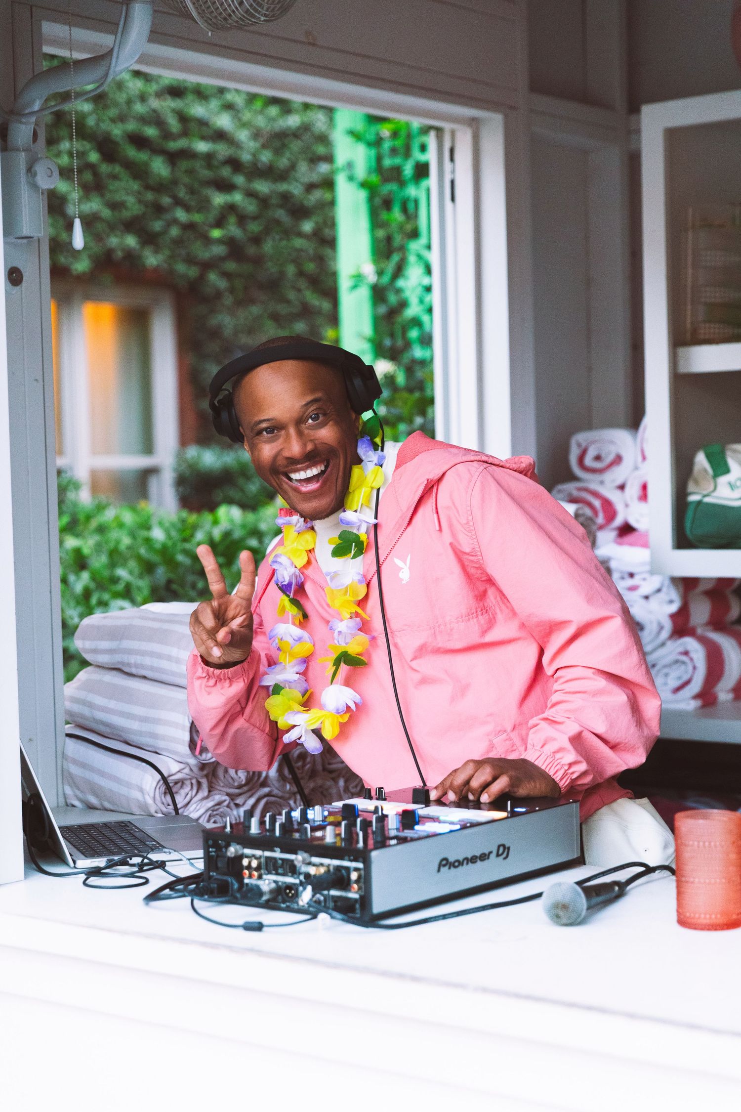 Smiling DJ in pink jacket and lei giving peace sign at Pioneer DJ mixer setup.