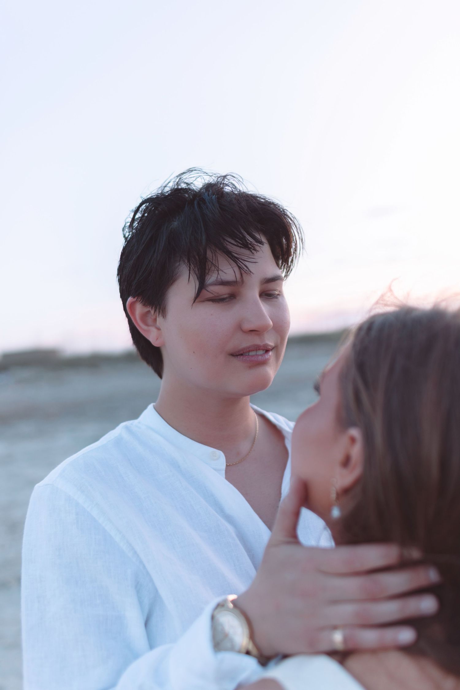 Two women sharing an intimate moment on a beach at sunset, one with short dark hair smiling.