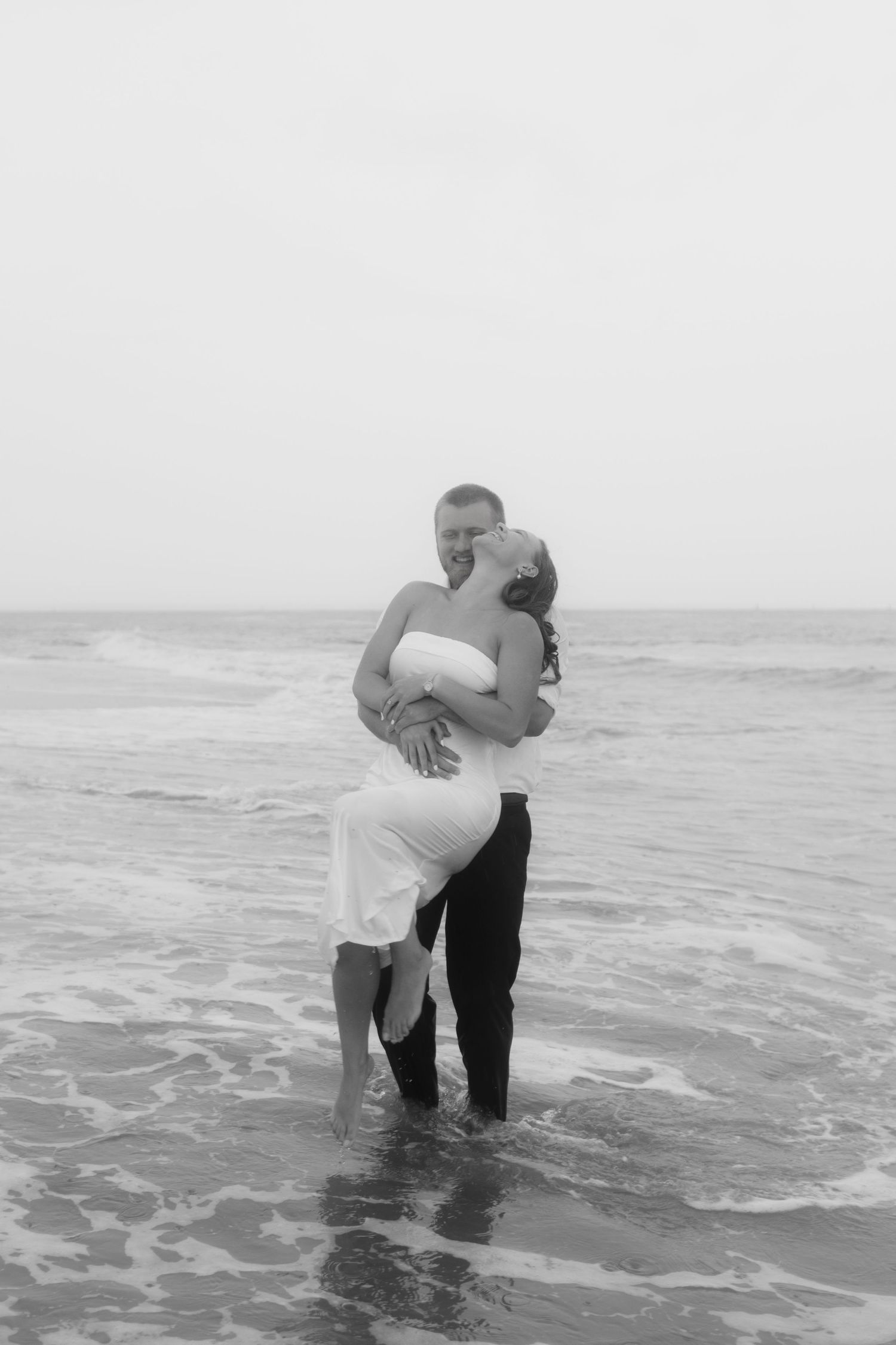 Black and white photo of a couple embracing in ocean waves, wearing white dress and black pants on a beach.
