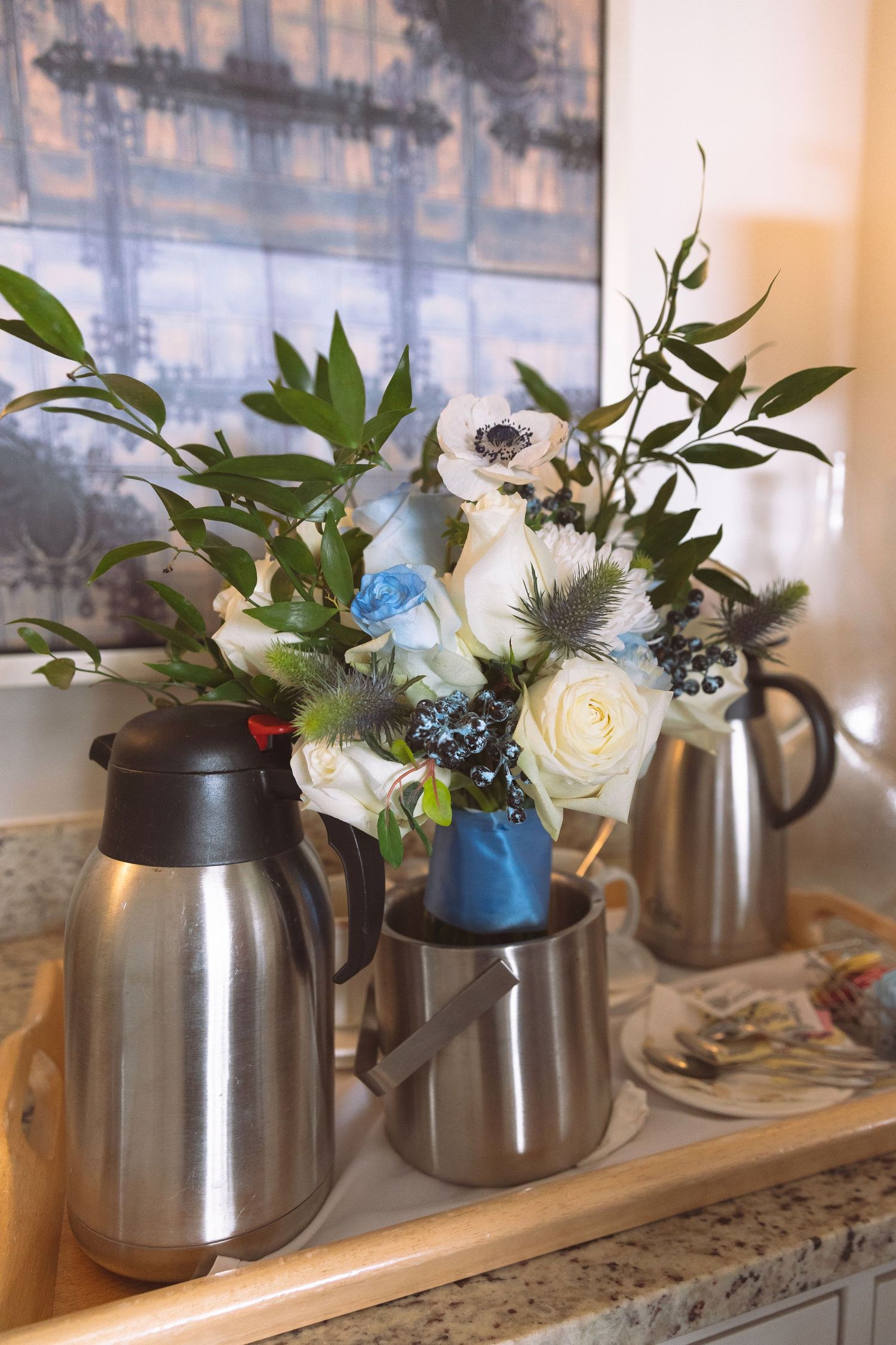 Silver coffee urns displayed beside white and blue floral arrangement on marble counter.