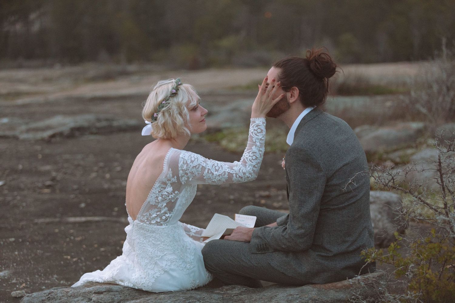 Bride tenderly touching grooms face while exchanging vows during an outdoor wedding ceremony on rocky terrain.