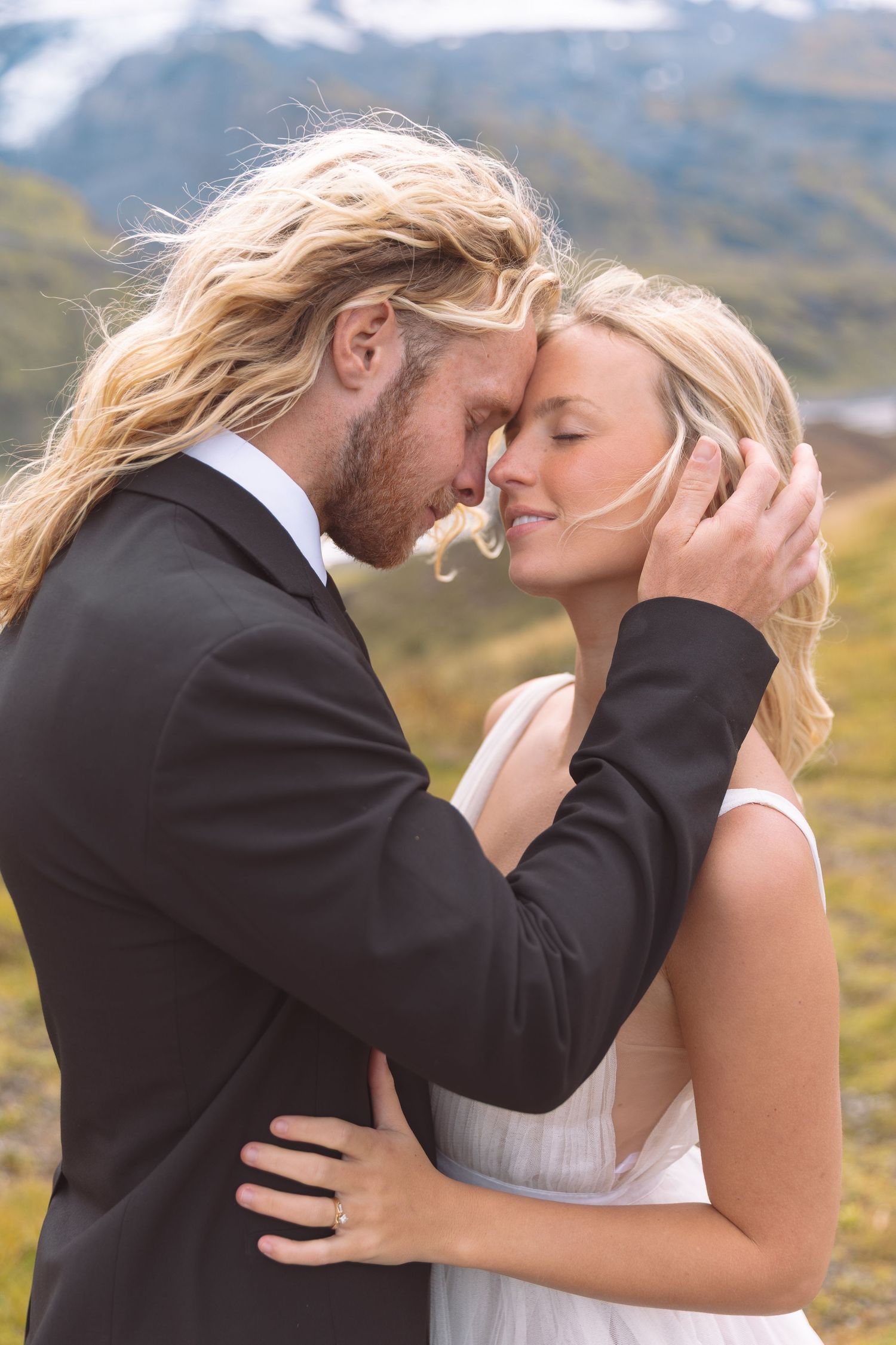 Groom tenderly cups brides face as they share an intimate moment with snowy mountains behind them.