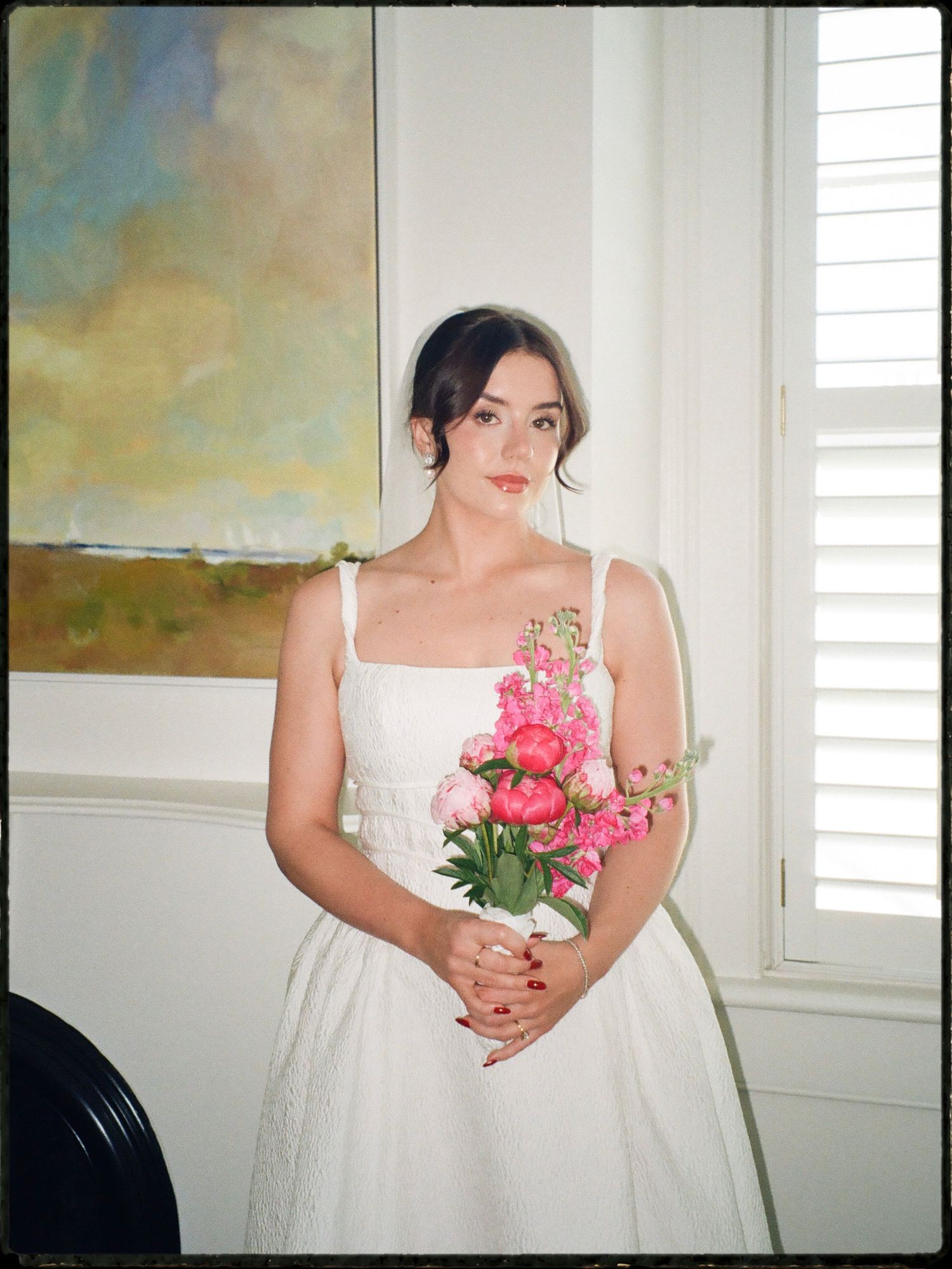Bride in white dress holding pink peony bouquet standing by window with abstract painting behind.