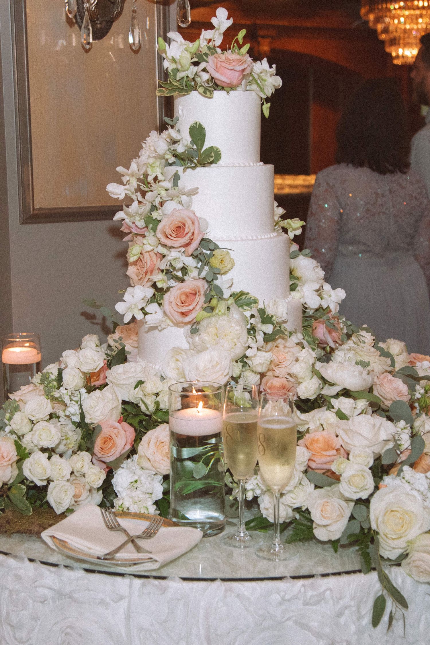 Elegant four-tier white wedding cake adorned with blush roses and champagne flutes on floral table.