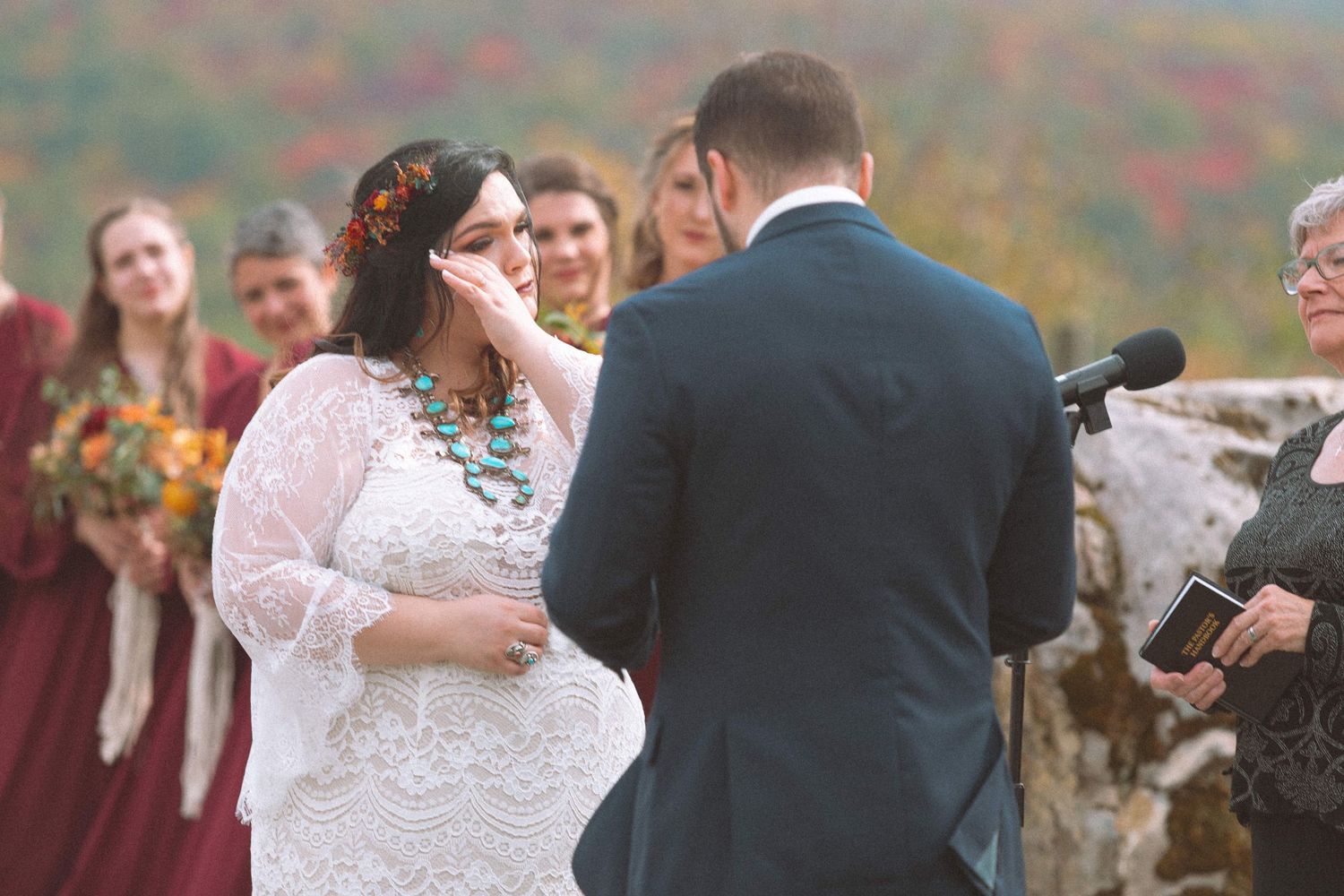 Emotional bride wiping tears during outdoor fall wedding ceremony with bridesmaids in burgundy.