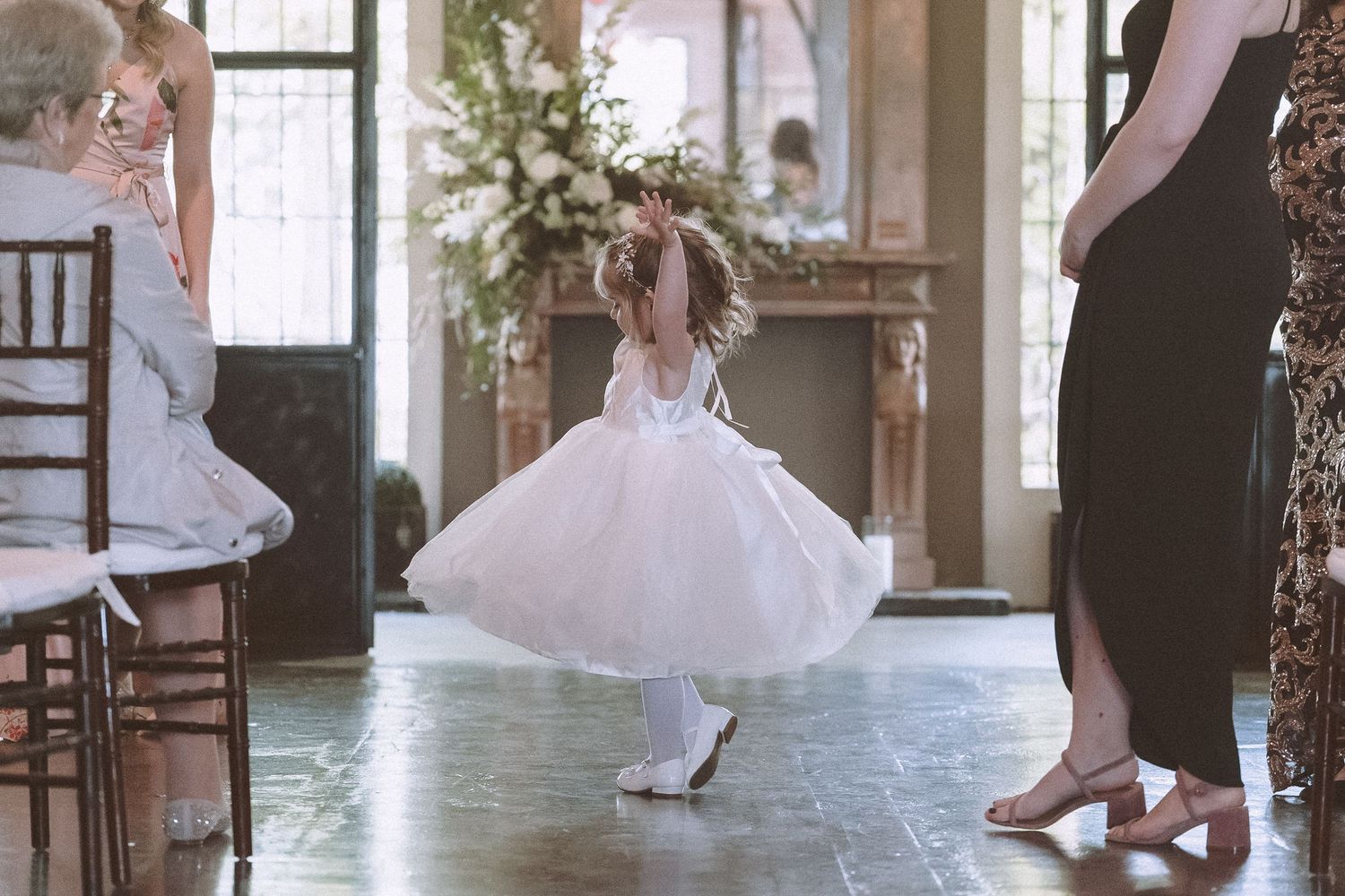 Flower girl in white tulle dress spinning joyfully during indoor wedding ceremony.