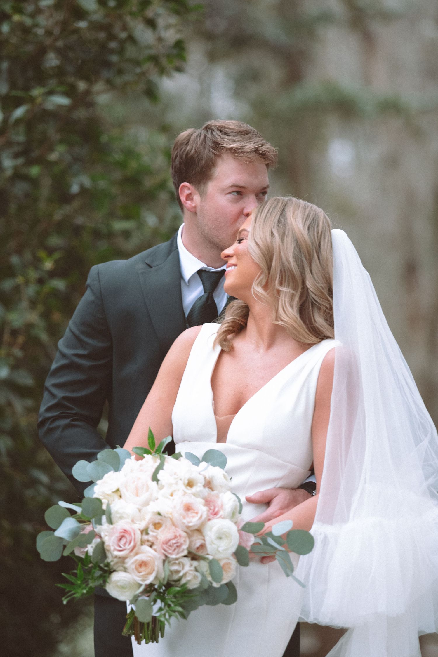 Groom kisses bride on cheek as she smiles holding blush pink and white floral bouquet.