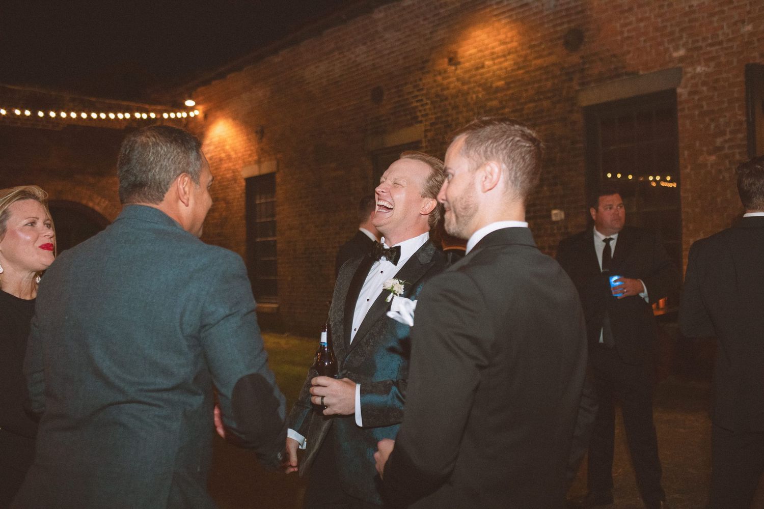 Wedding guests laughing and socializing outdoors at a nighttime reception at a rustic brick venue.