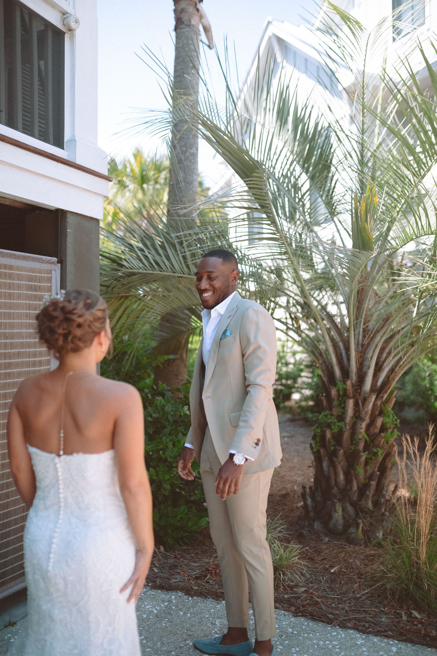 Groom in tan suit smiling at bride during outdoor first look moment surrounded by tropical palms.